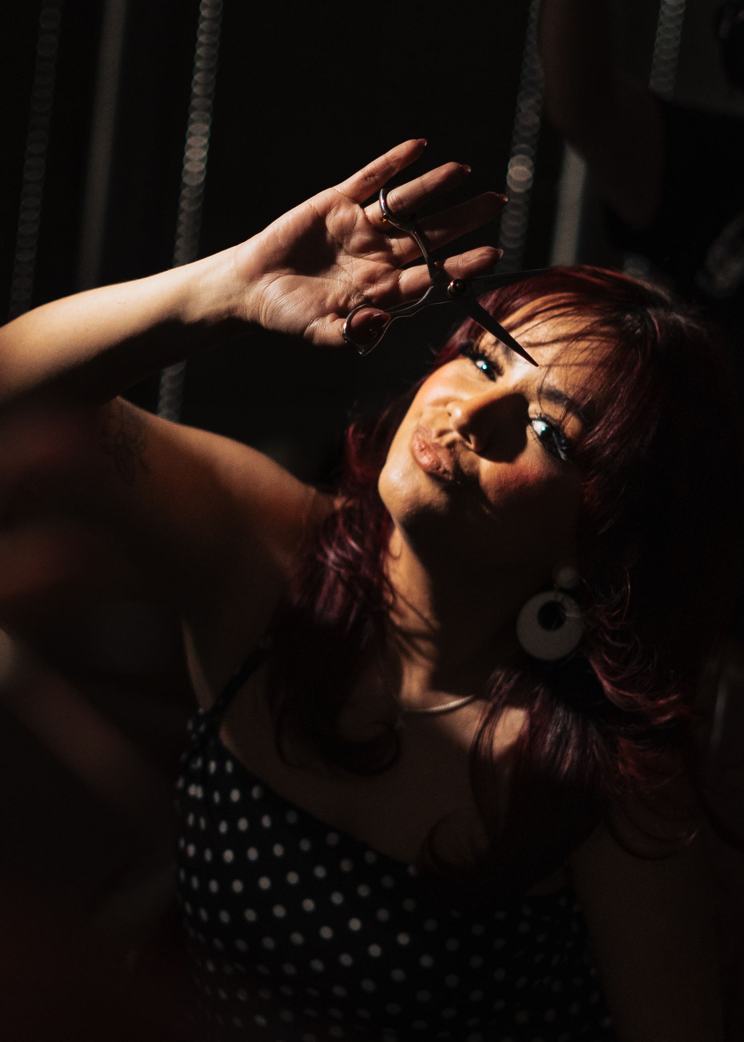 Woman with red hair and round earrings holding scissors up dramatically in dramatic moody salon lighting.