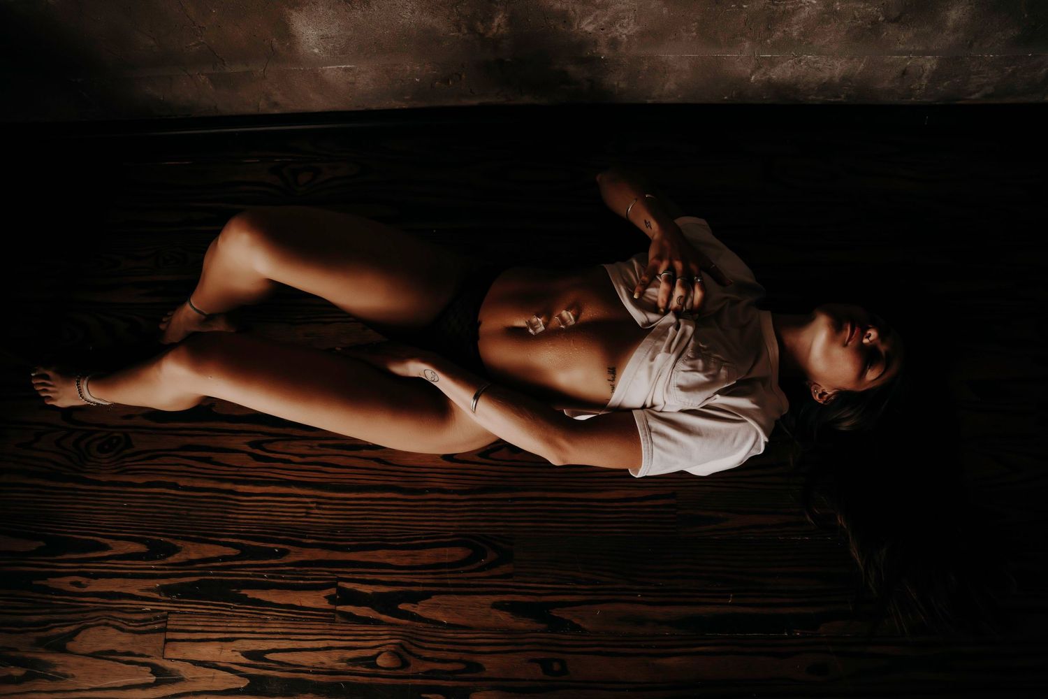 Woman in white shirt lying on wooden floor in moody low-light boudoir portrait.