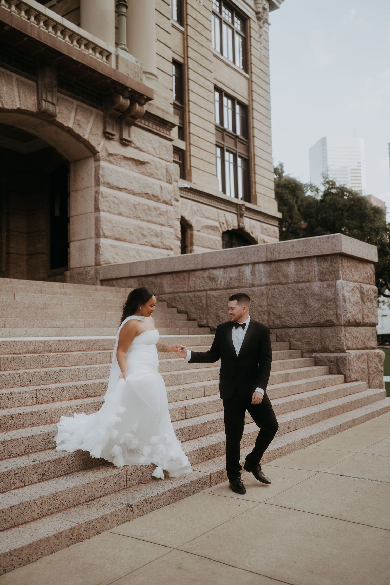 Bride in ruffled white gown and groom in black tuxedo walk hand-in-hand up stone steps of historic building.