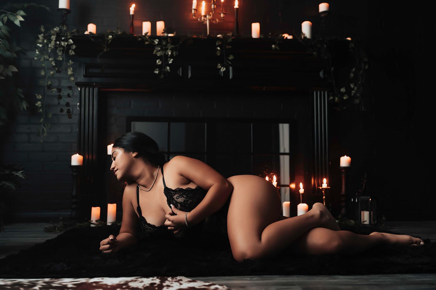 Woman in black lingerie posing by candlelit fireplace in dramatic dark boudoir setting.