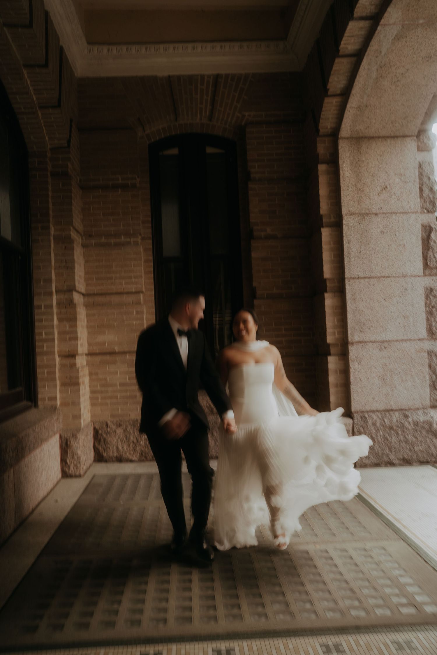 Blurred motion shot of laughing bride and groom walking through historic building archway.