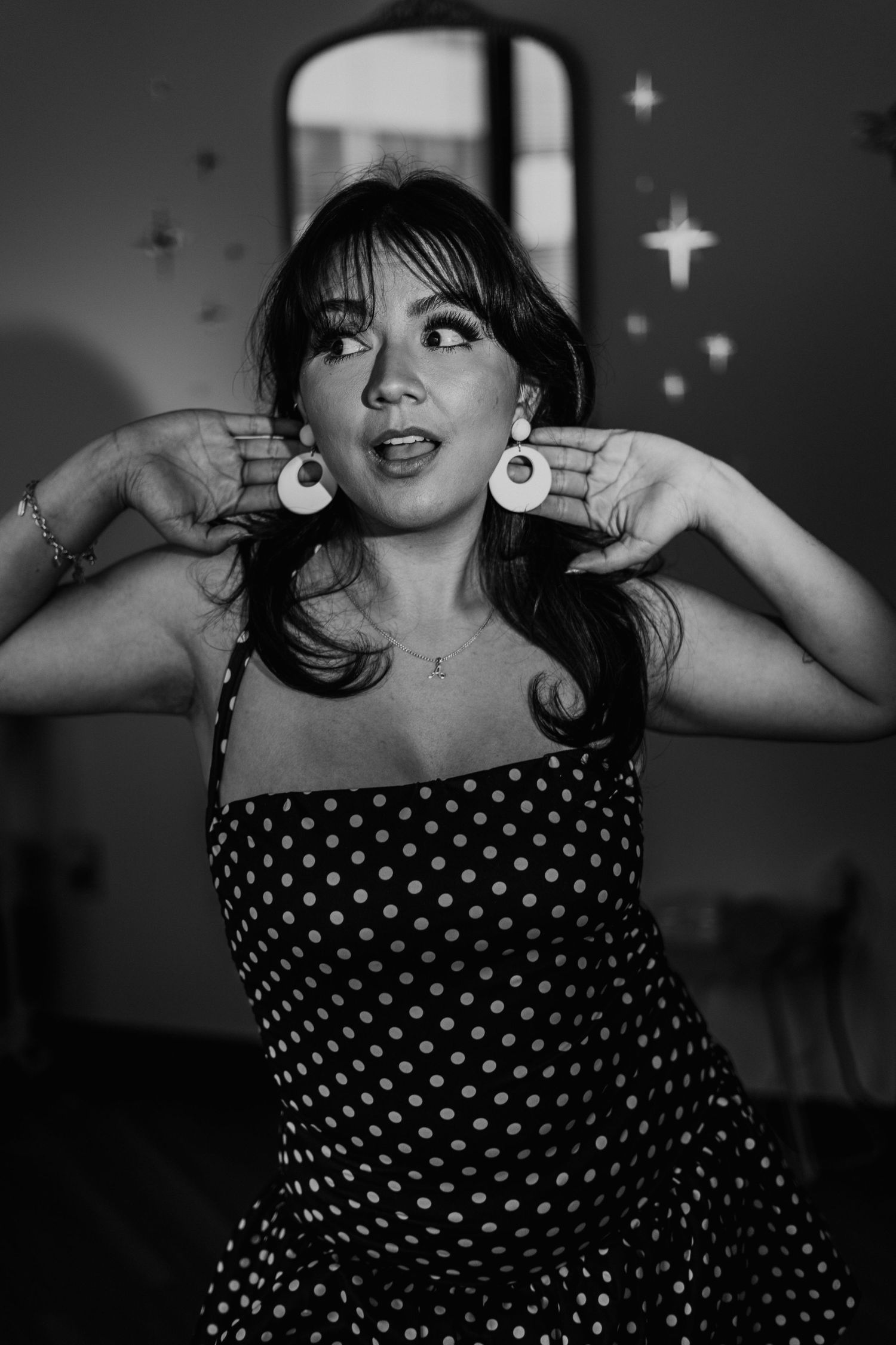 Black and white photo of a young woman in a polka dot dress pointing to her large circular earrings.