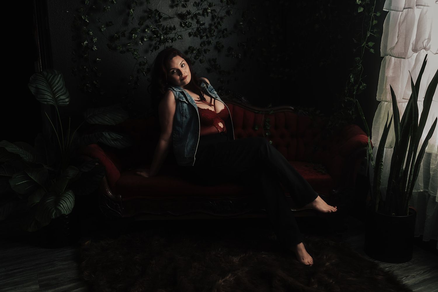 Woman in denim vest and black jeans lounging on red velvet sofa in dark moody interior setting.