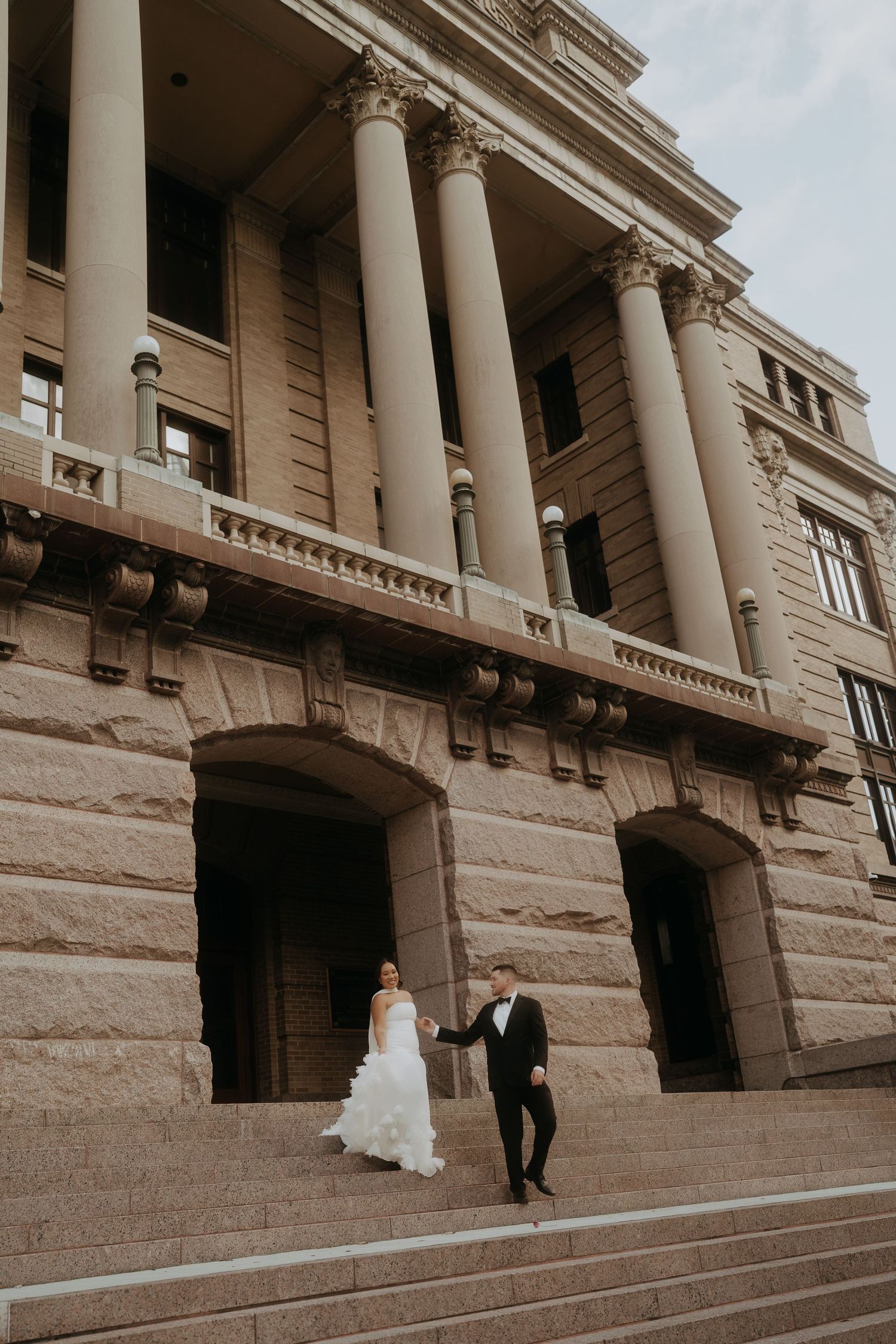 Bride and groom descend grand stone steps of a neoclassical building with towering columns together.