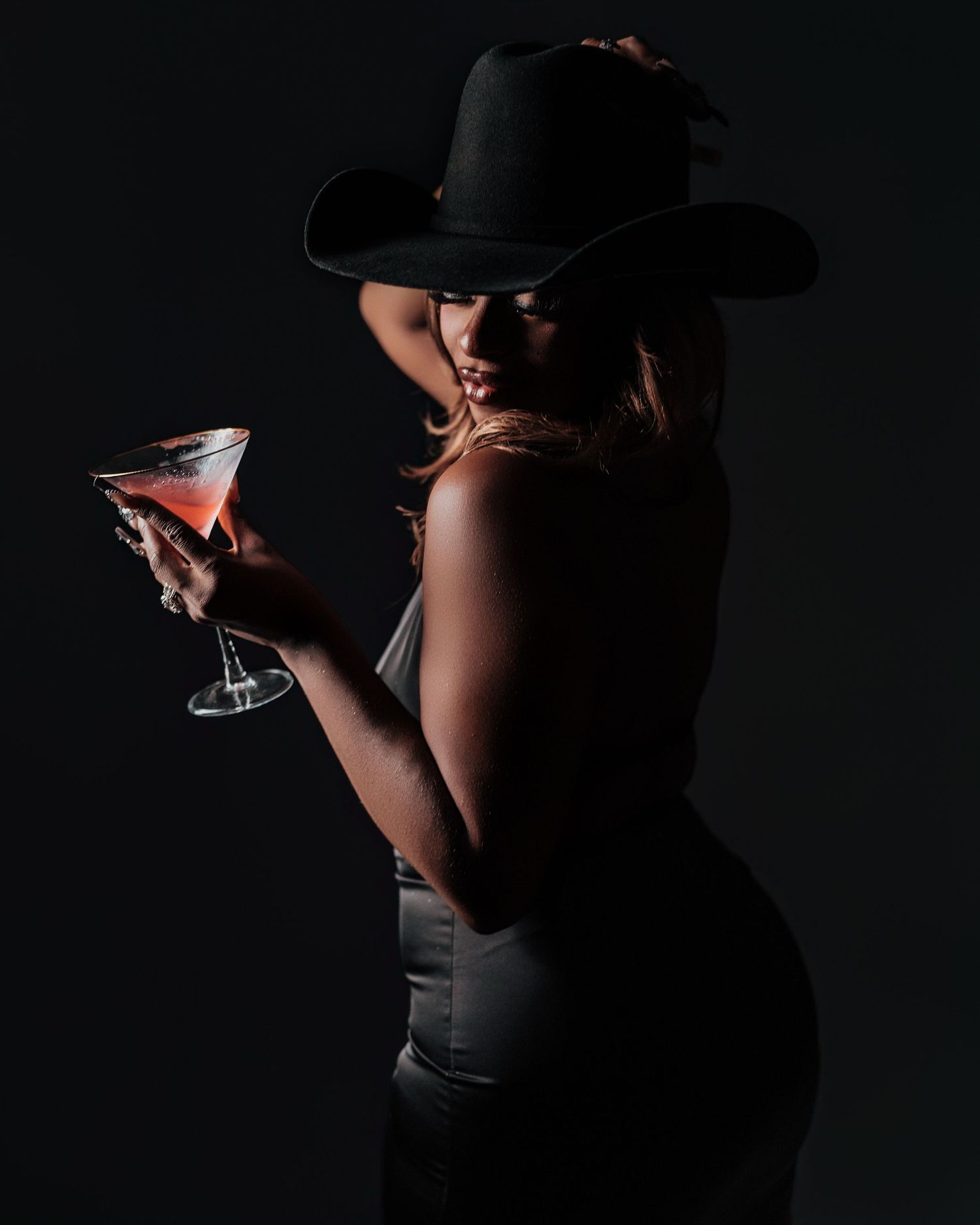 Silhouetted woman in black cowboy hat holds cocktail glass in dramatic dark studio lighting.