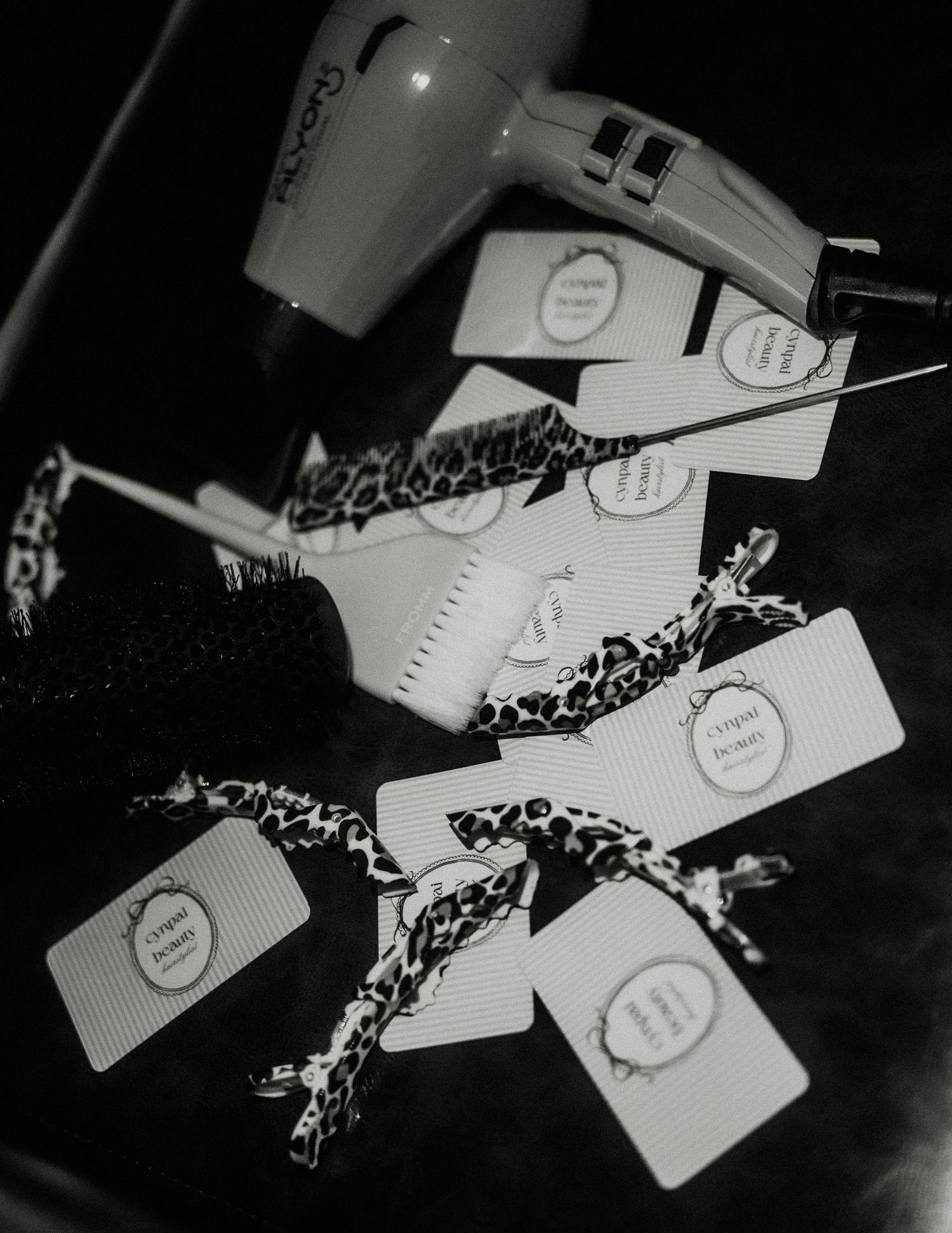 Black and white flat lay of hair dryer, round brush, and gift tags with leopard print ribbons on leather.