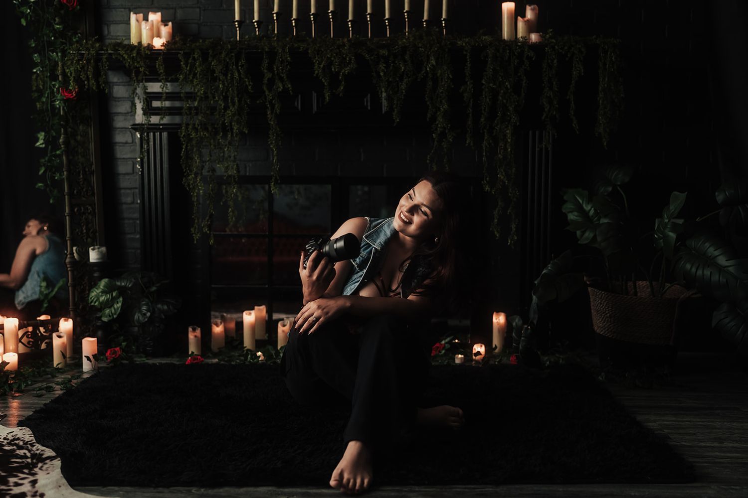 Smiling woman relaxing on dark rug with candles and lush greenery near fireplace in moody atmospheric space.