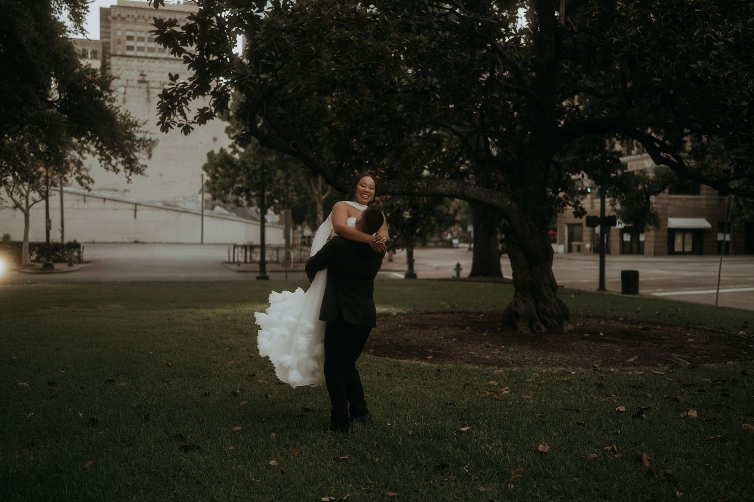 Groom lifts bride in white wedding gown as she laughs in an urban park at dusk.