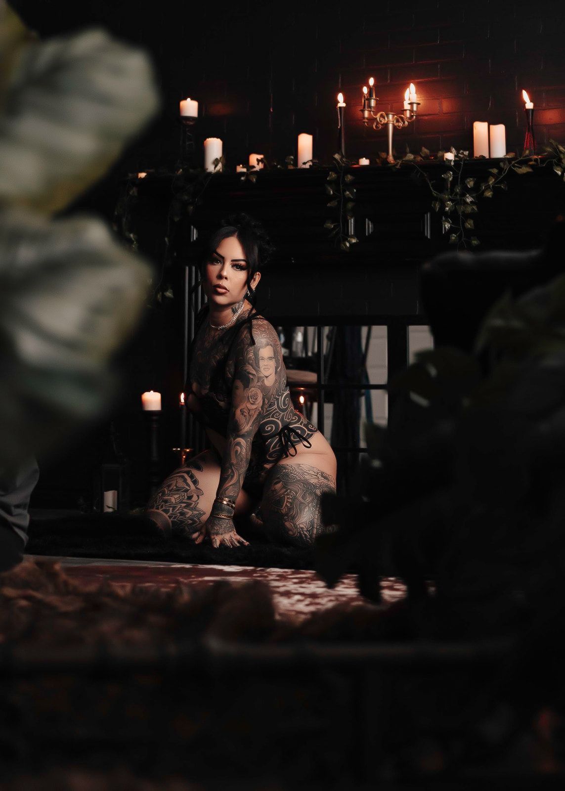 Heavily tattooed woman posing in candlelit dark room surrounded by candles and greenery in artistic photography.