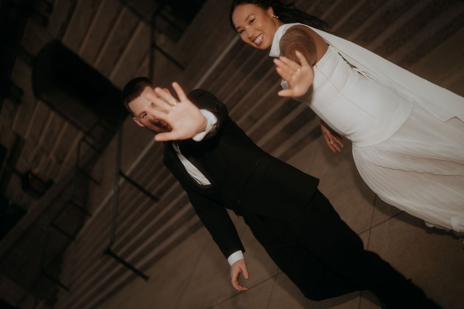 Bride and groom dancing playfully with hands outstretched toward camera at wedding reception.