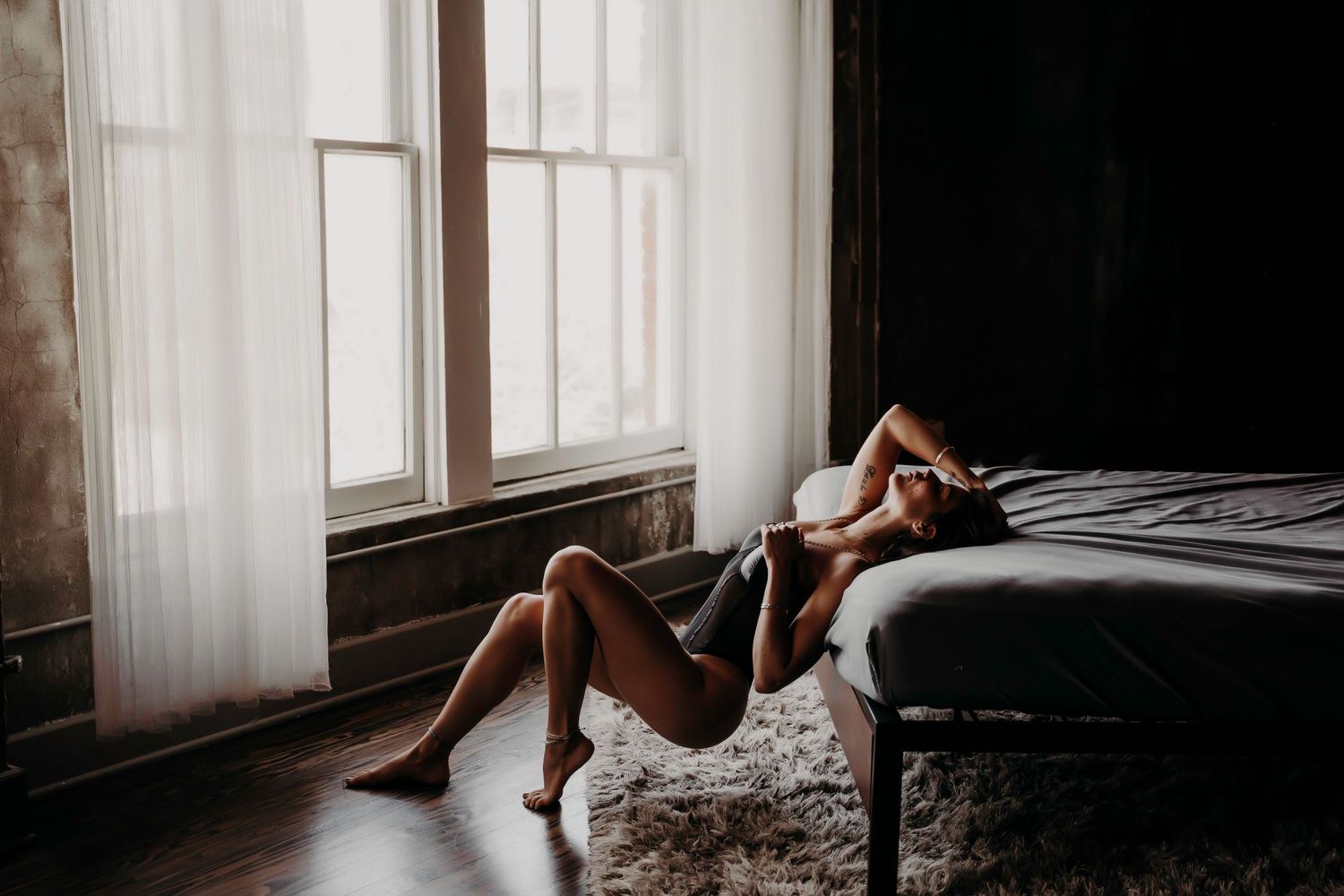 Woman in dark lingerie lying on floor beside bed in moody boudoir portrait.