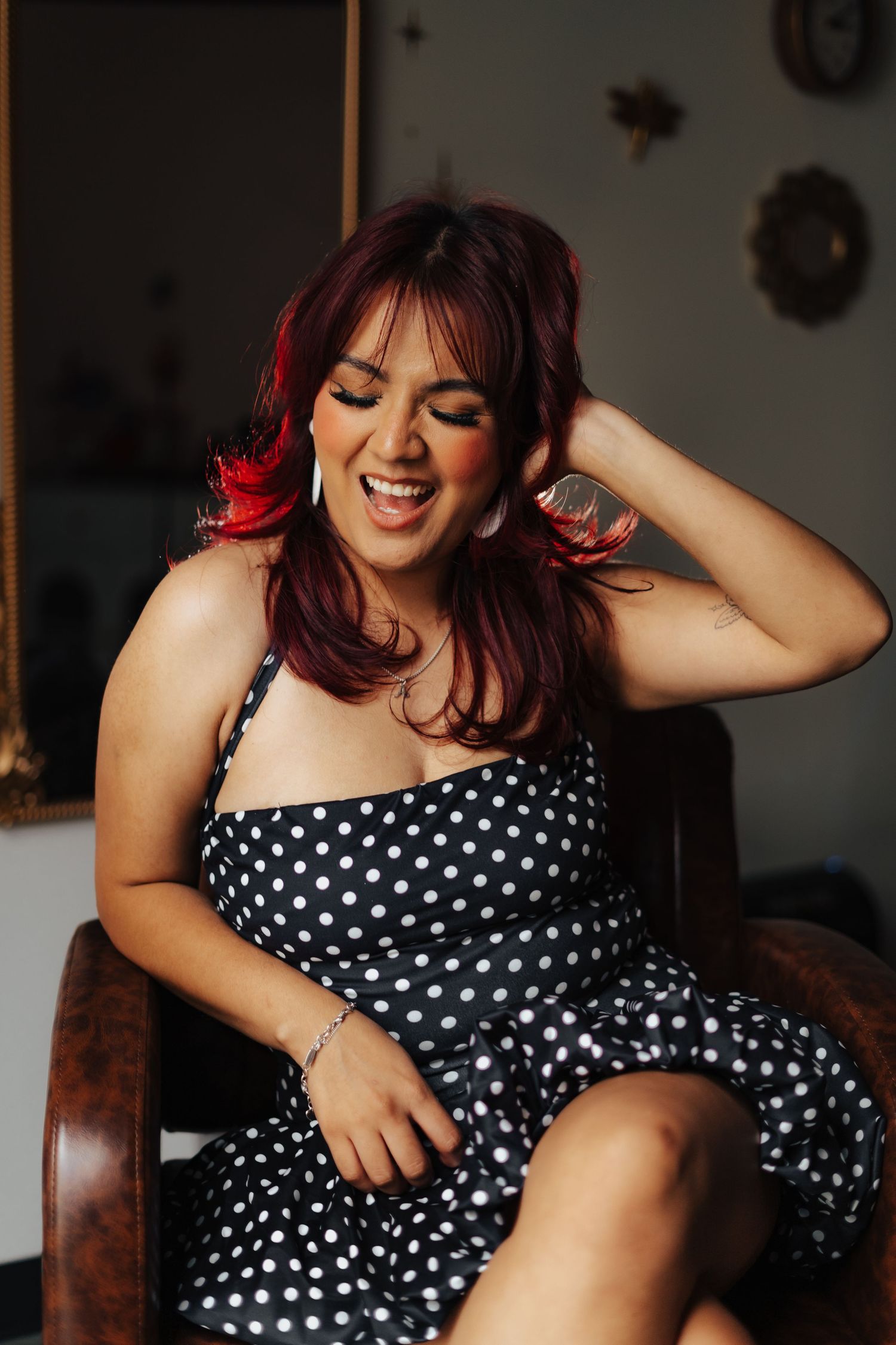 Smiling red-haired woman in a black polka dot dress laughing joyfully while seated in a chair.