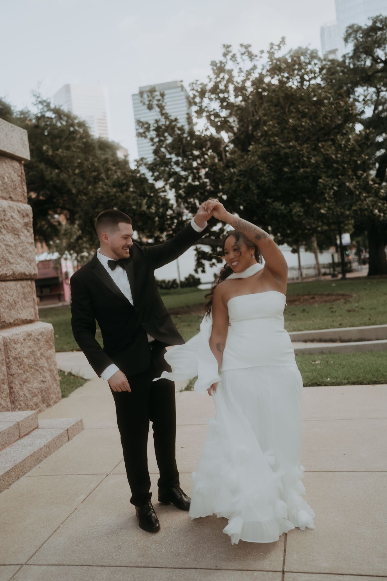 Joyful groom twirls smiling bride in white ruffled wedding dress outdoors with city skyline in background.