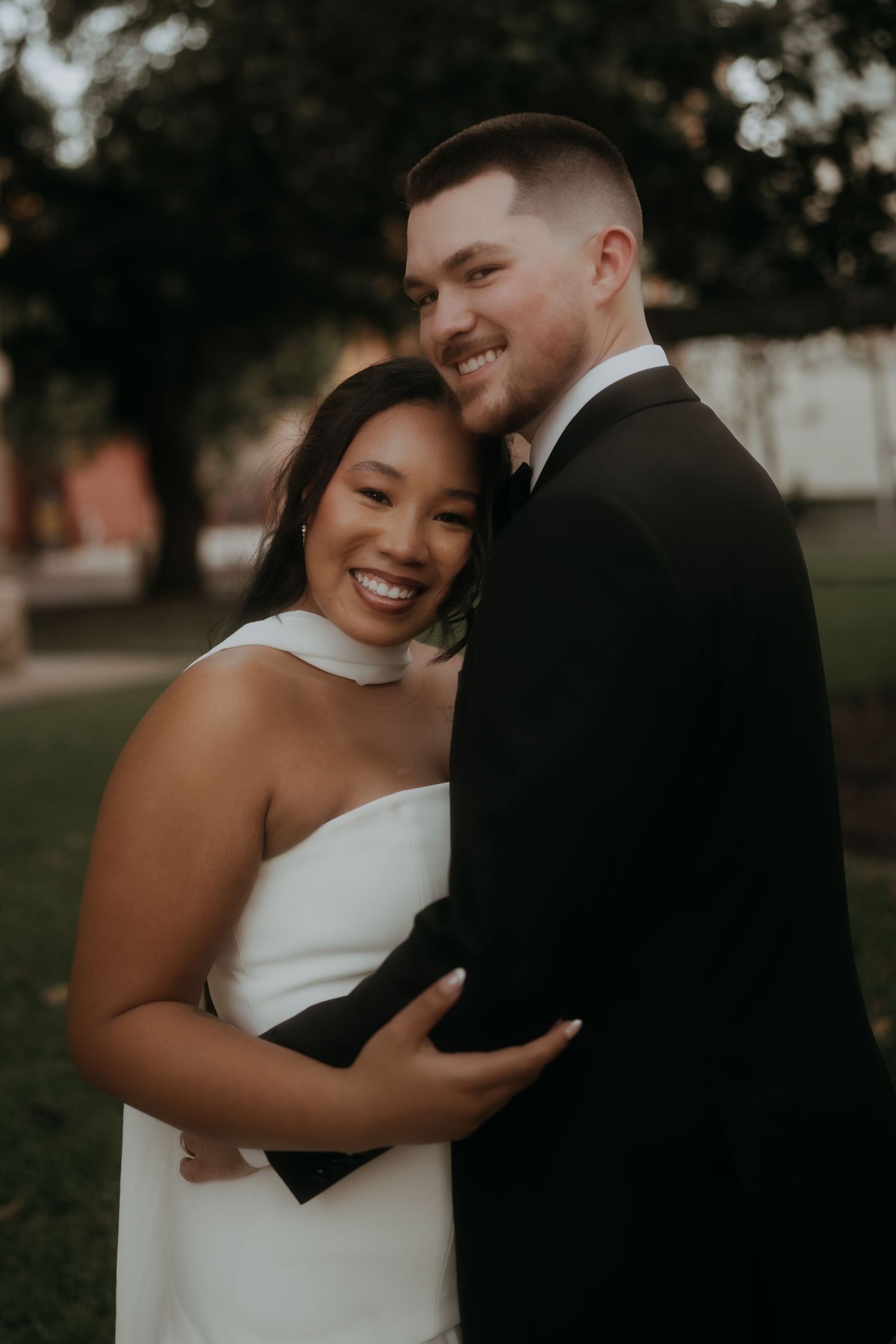 Bride in white strapless gown embraces groom in black tuxedo outdoors during golden hour wedding portraits.
