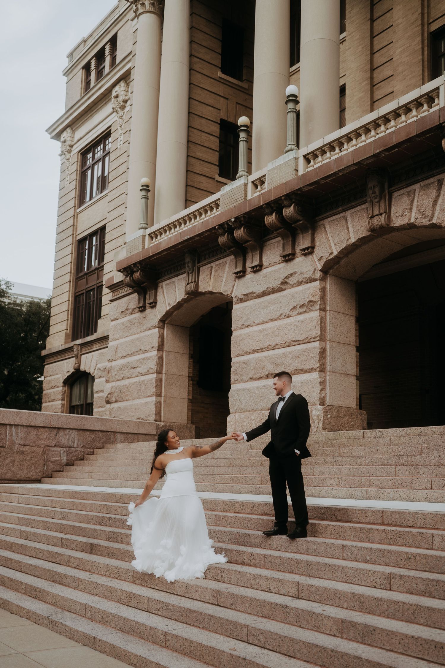 Bride and groom hold hands on grand stone steps of historic building on their wedding day.
