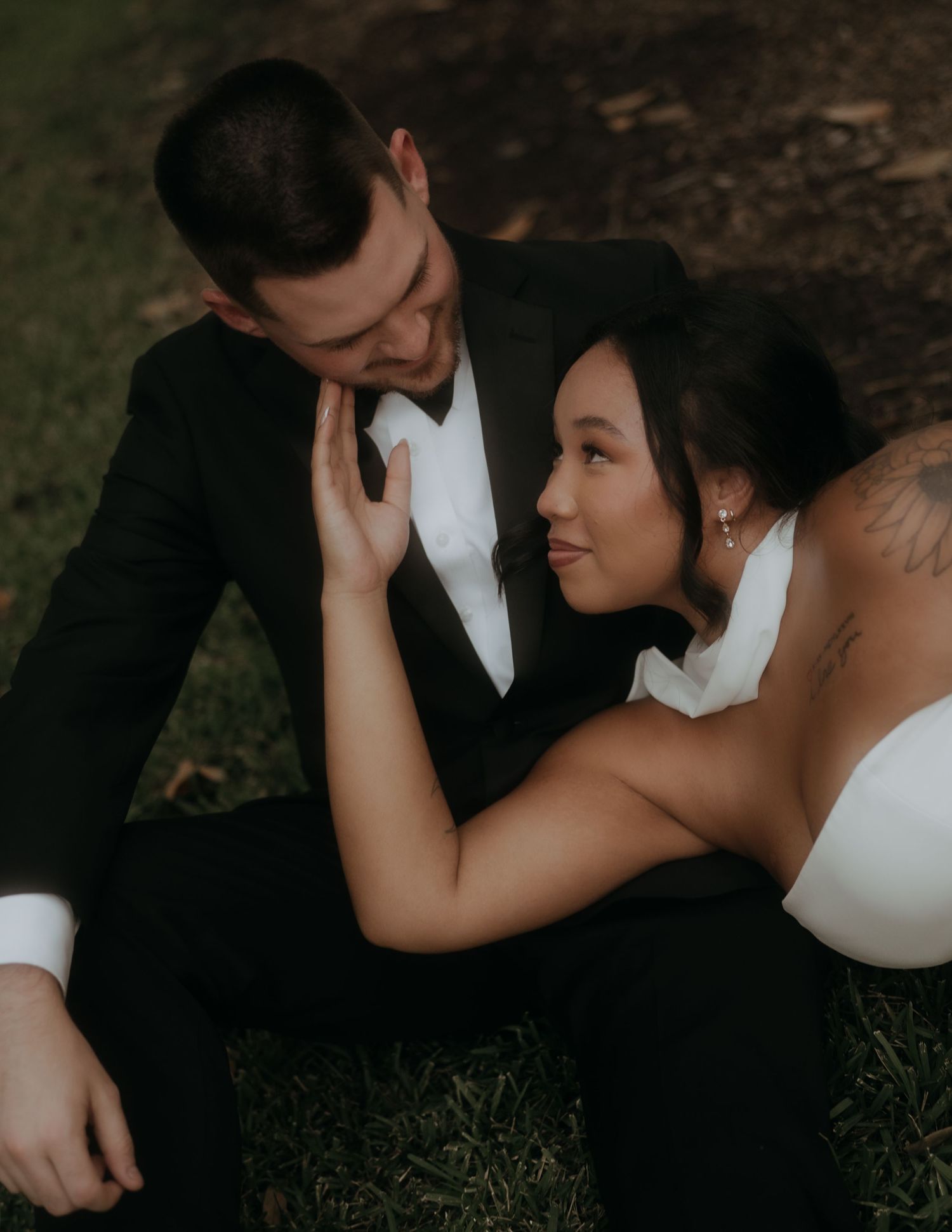 Bride in white halter gown gazes at groom in black tuxedo while sitting together on grass.
