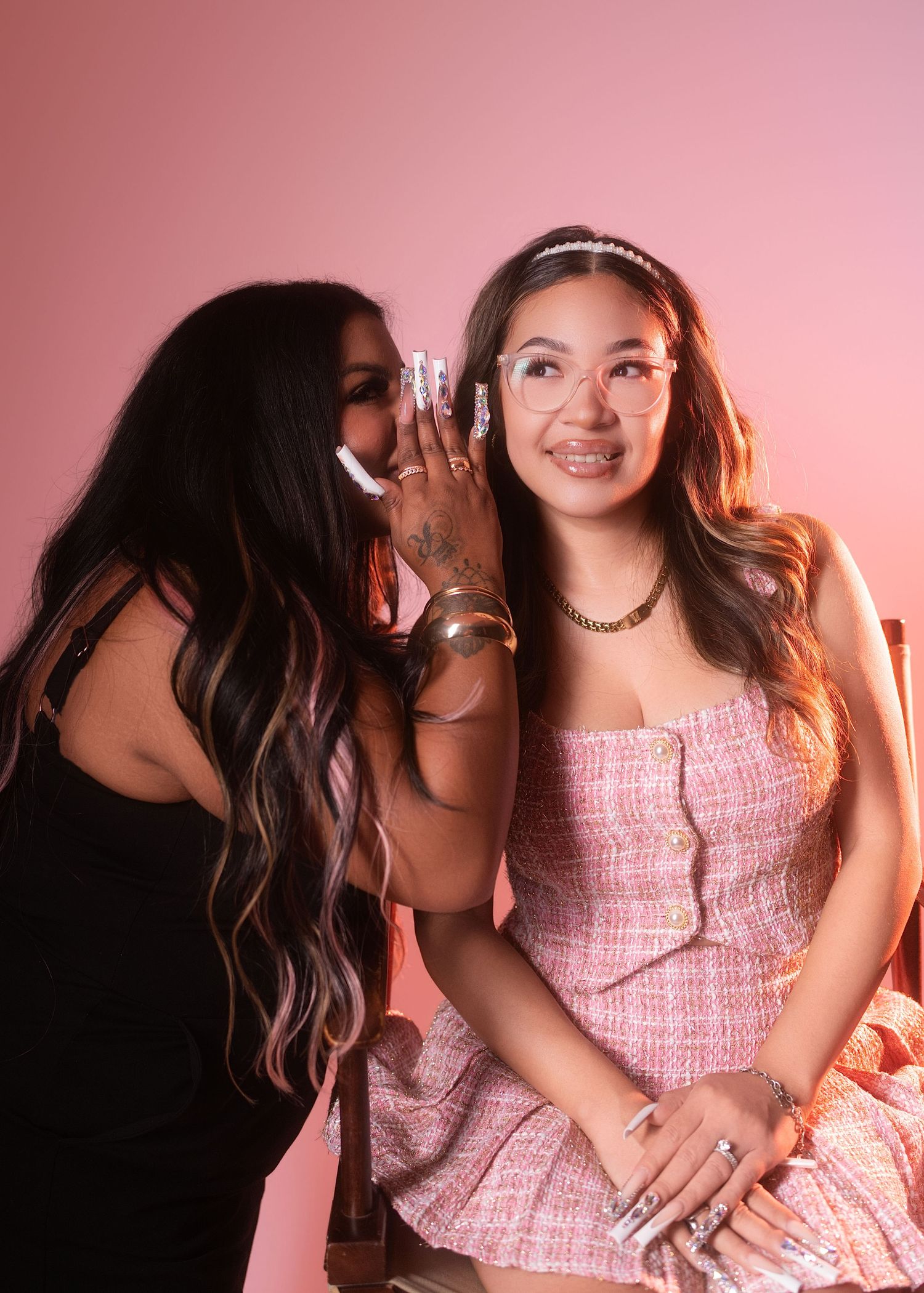 Two young women sharing a secret, one whispering into the other's ear in pink studio.