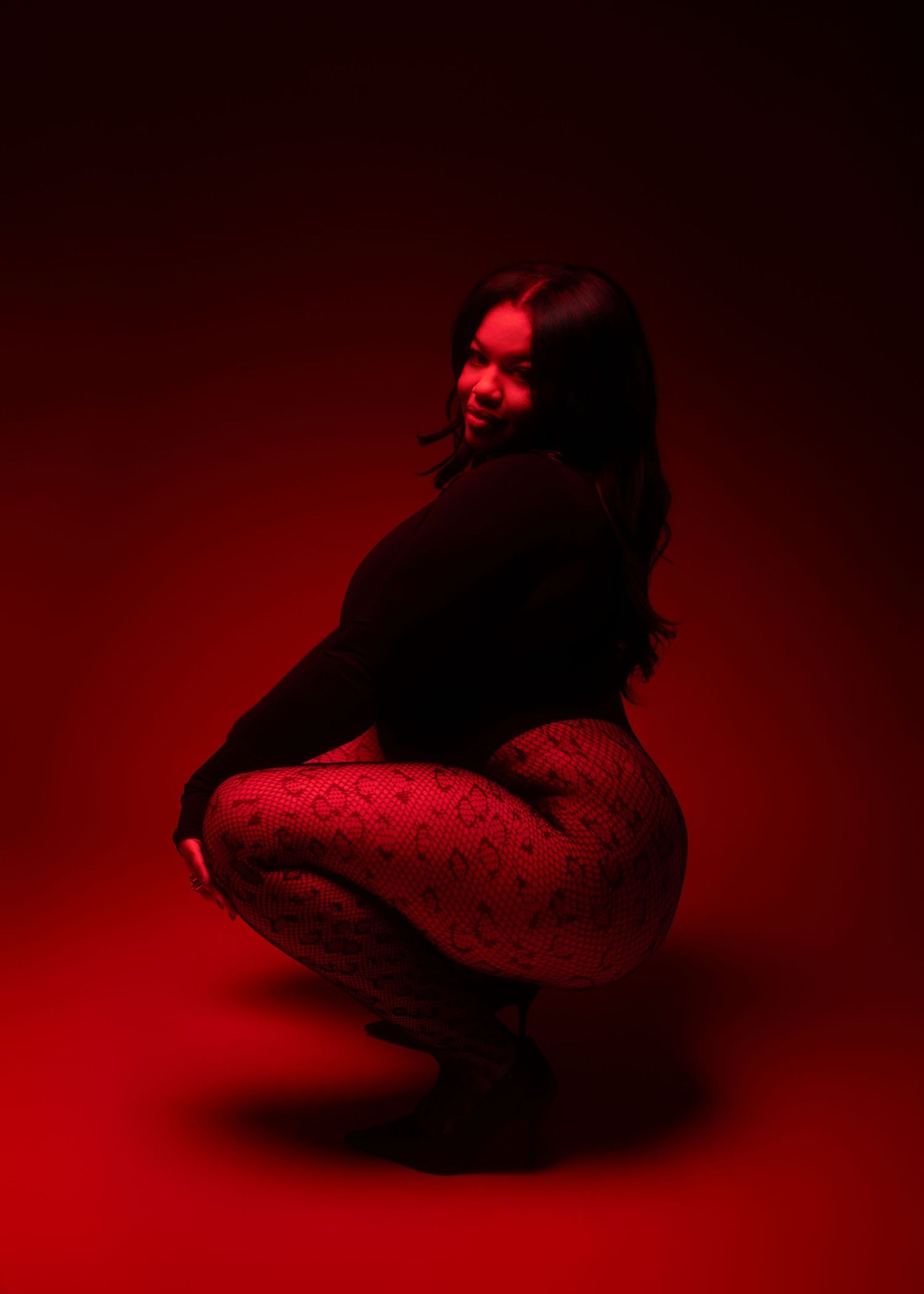 Woman crouching in red light wearing black top and patterned fishnet tights.
