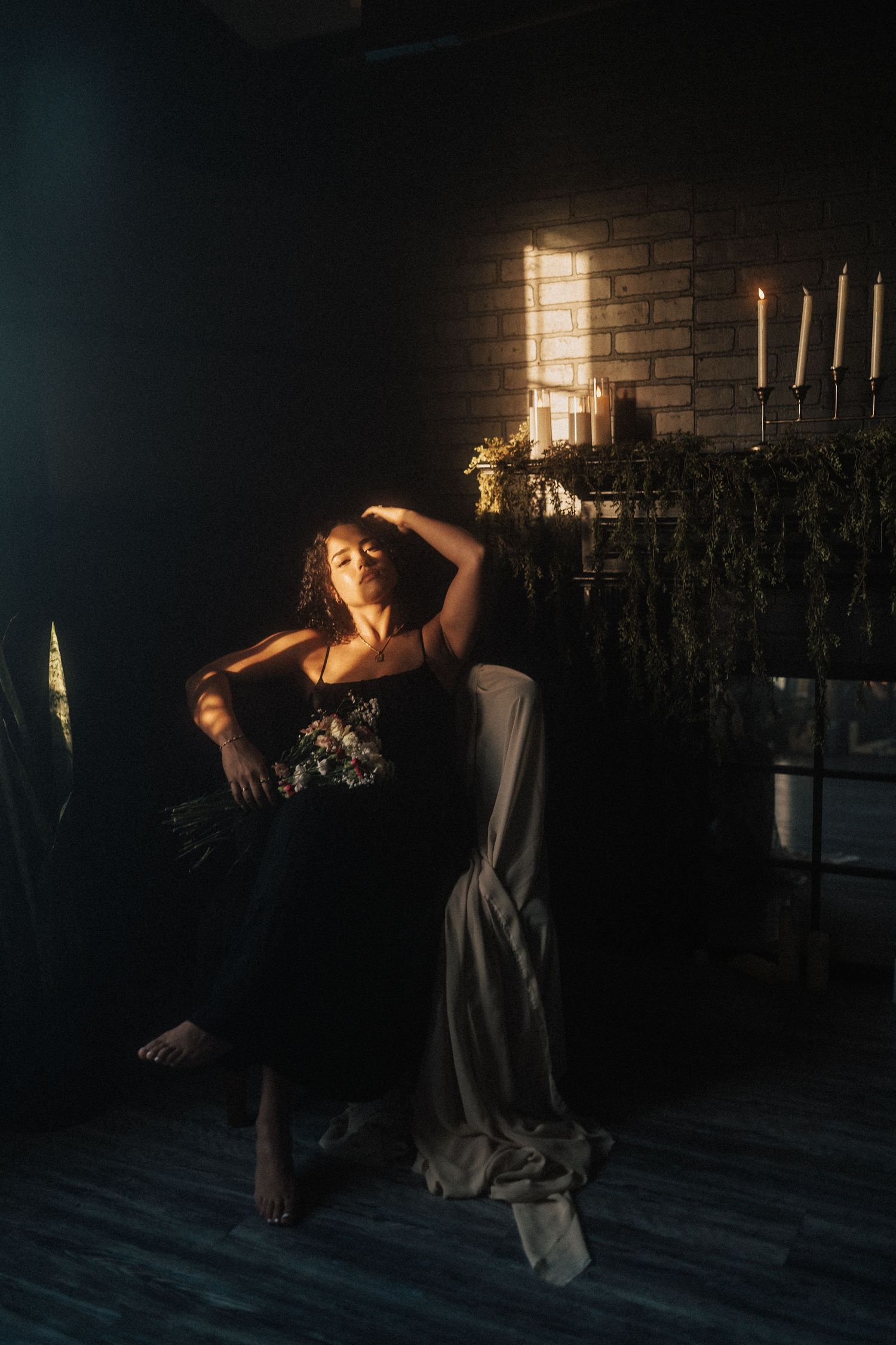 Woman in black floral dress reclining in chair with dramatic shaft of light and candles in dark studio.