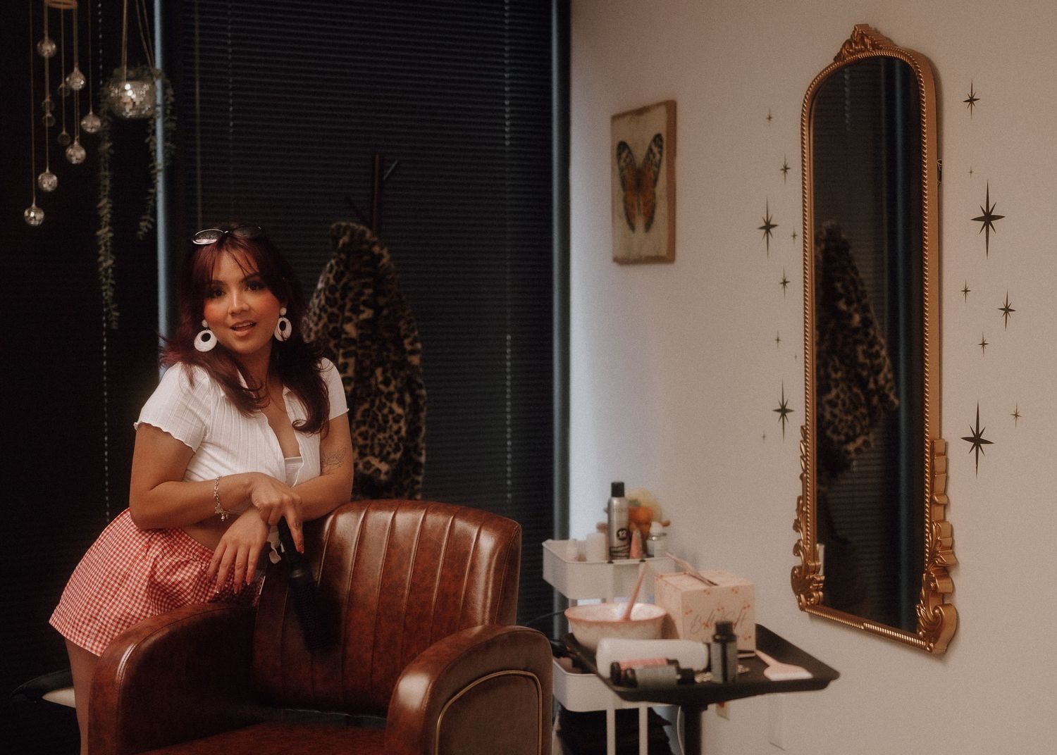 Smiling woman in pink skirt sitting in leather salon chair beside gold ornate mirror with star wall decor.