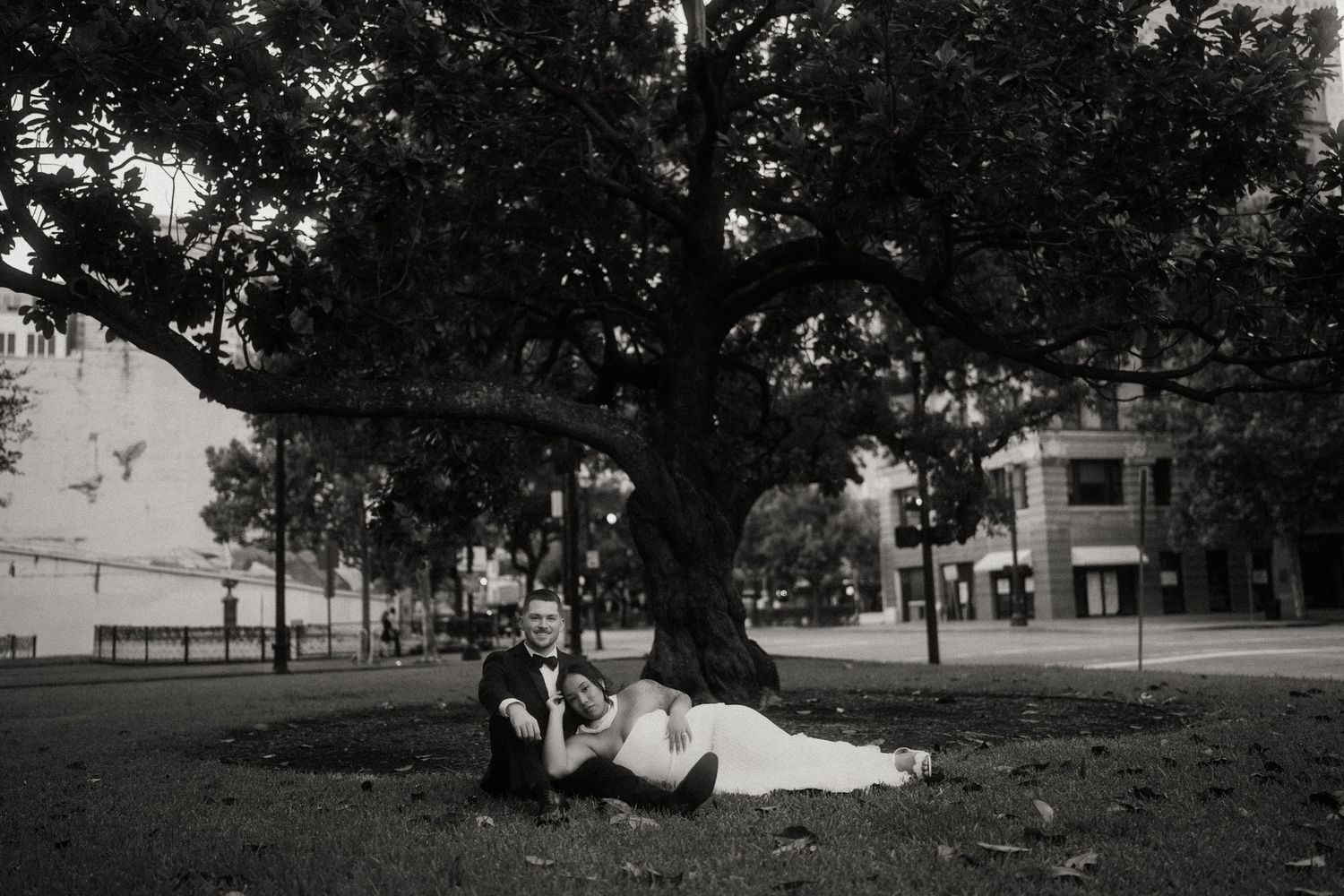 Black and white photo of bride and groom relaxing under a large tree in an urban park.