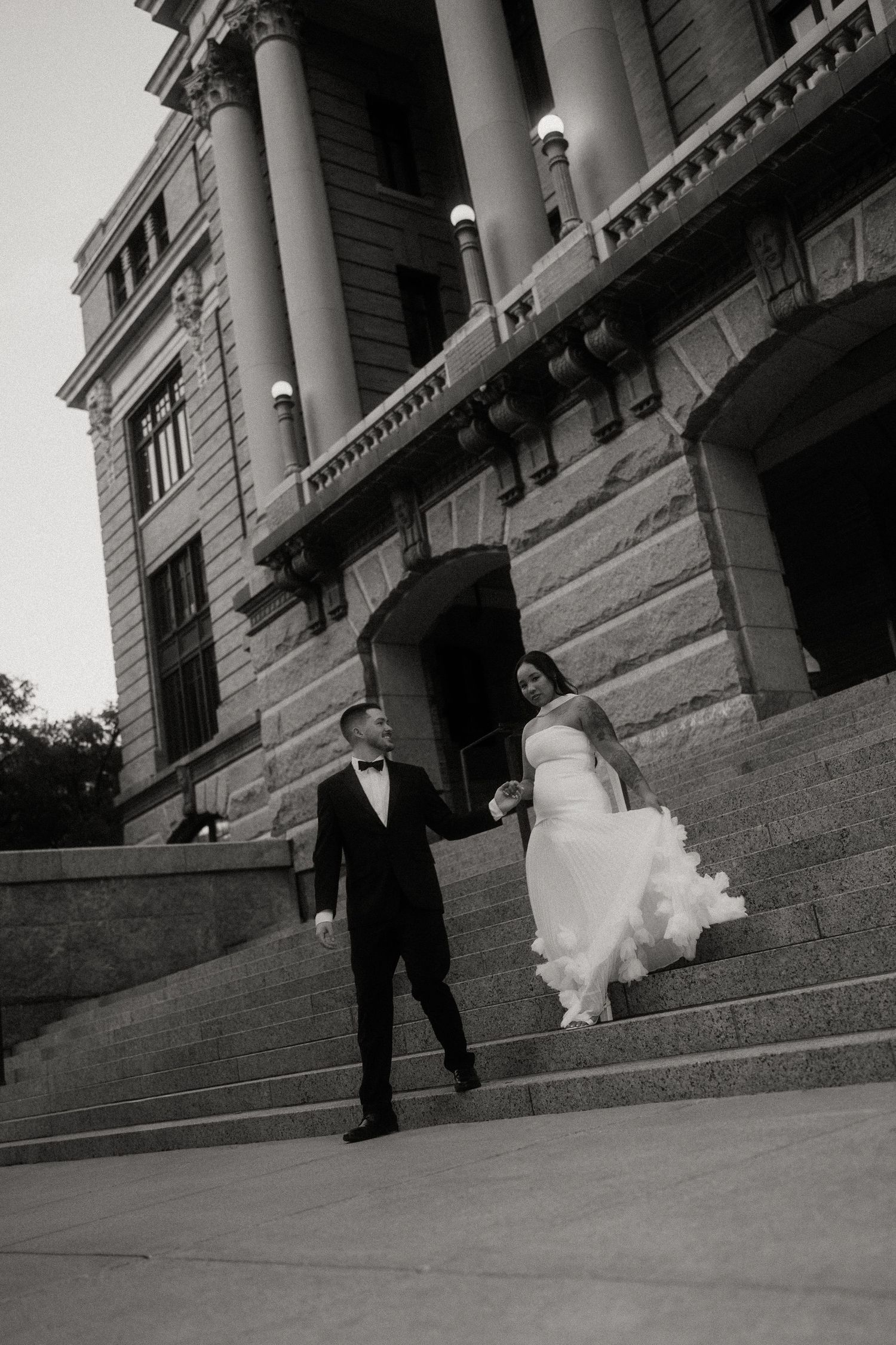 Black and white photo of bride and groom walking down steps of grand historic building.