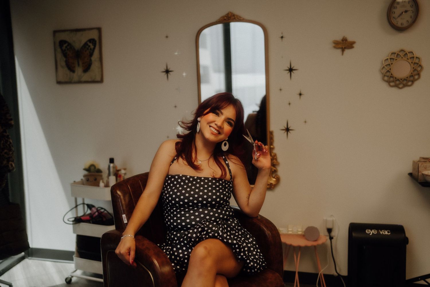 Smiling woman in black polka dot dress sitting in leather salon chair with gold mirror and star wall decor.