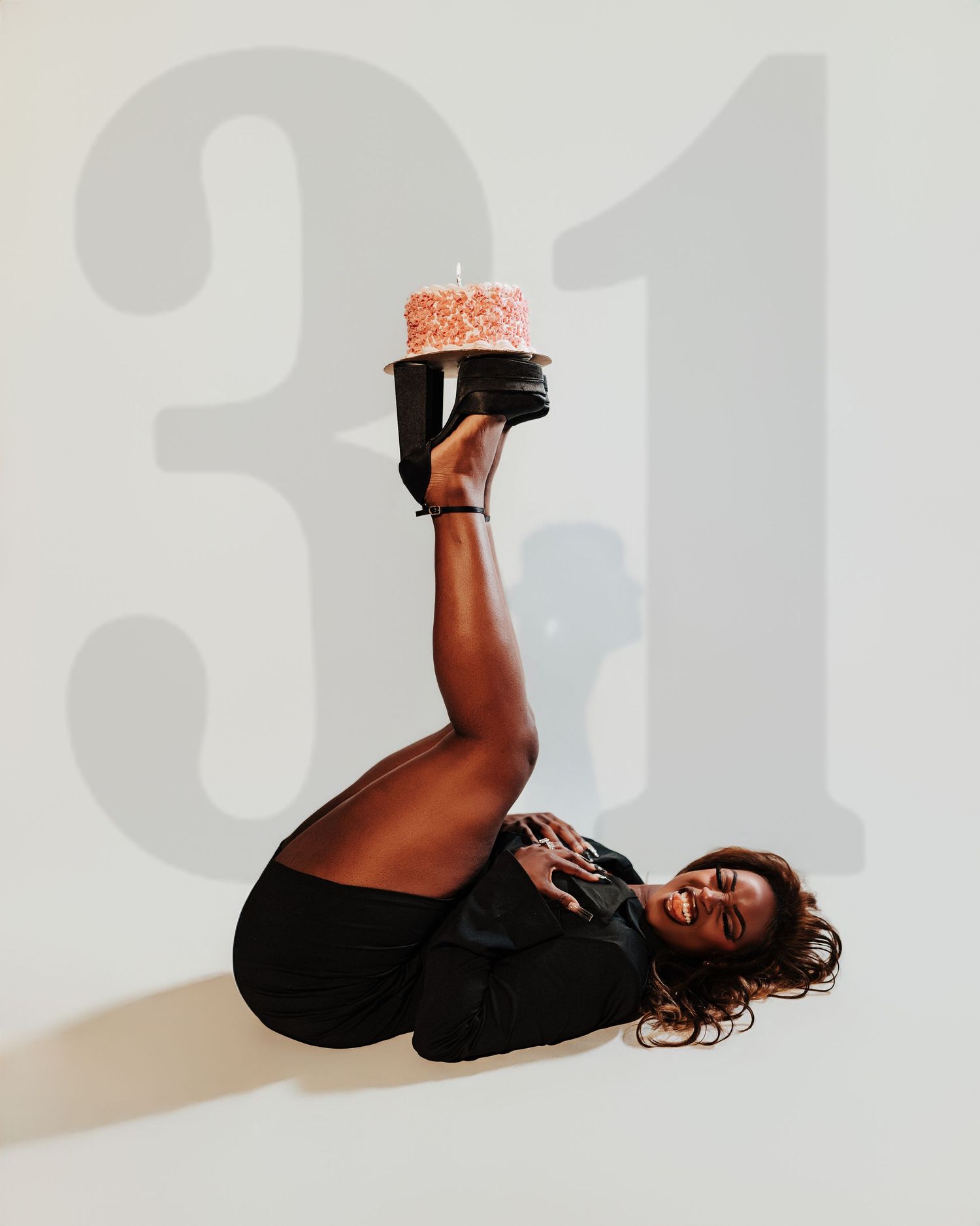 Creative birthday photo with woman balancing cake on heels beside large number 31 backdrop.