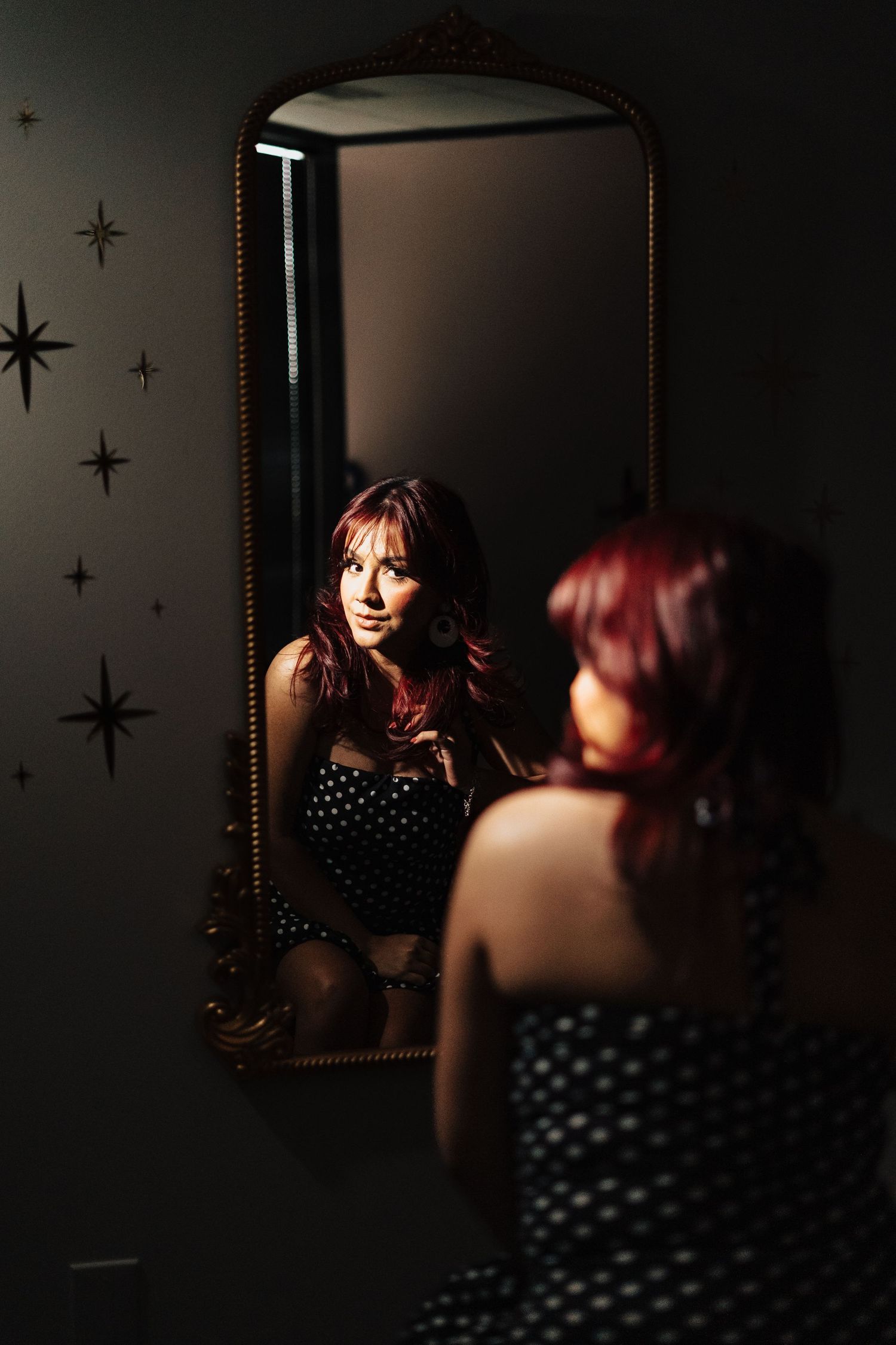 Woman with red hair in polka dot dress gazing at her reflection in an ornate gold mirror with star decor.