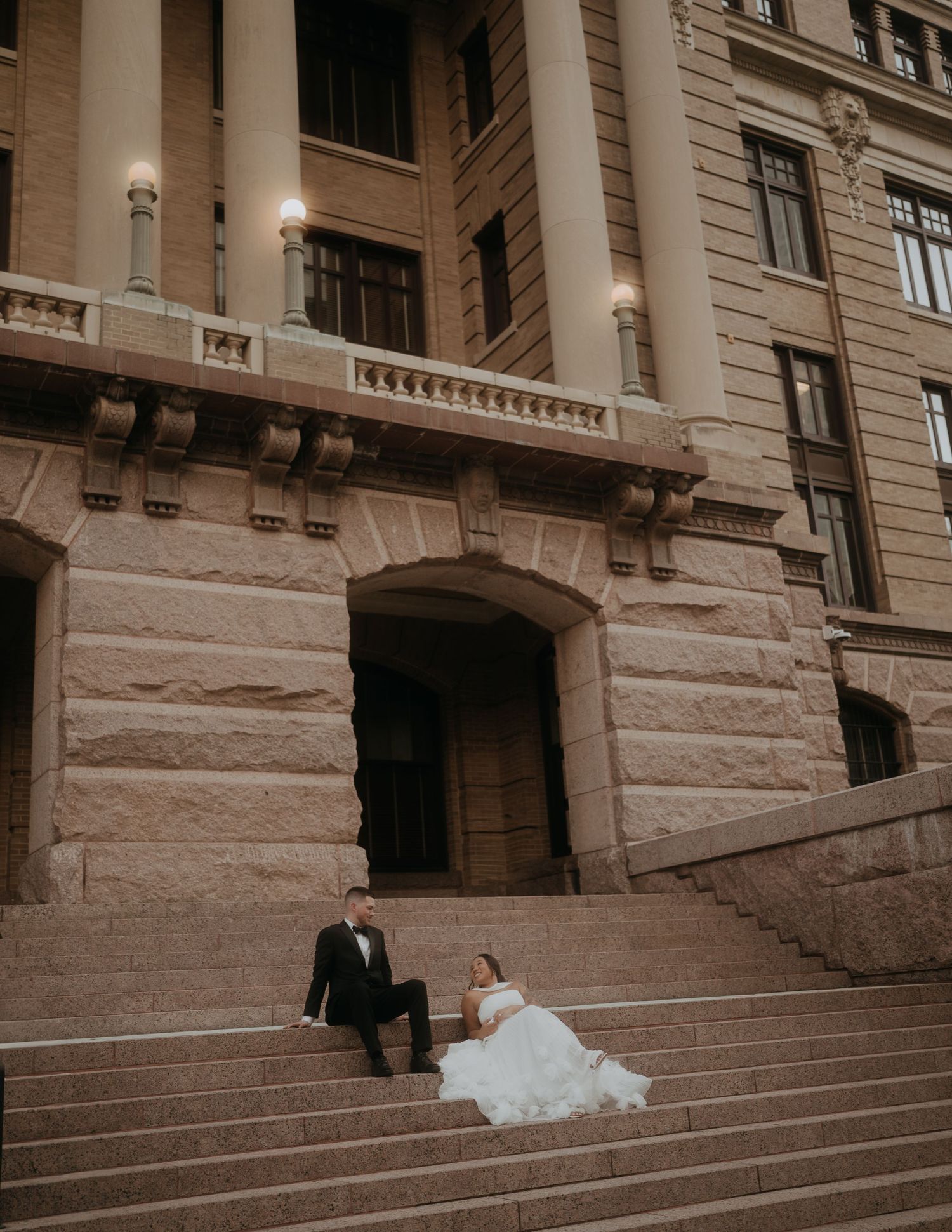 Wedding couple posing on grand stone steps outside elegant historic building at dusk.