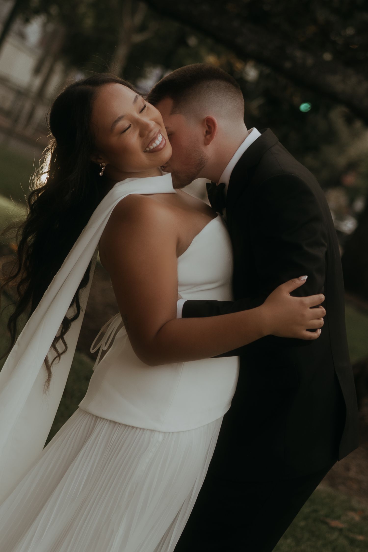 Groom in black tuxedo kisses bride on cheek as she smiles during outdoor wedding portraits.