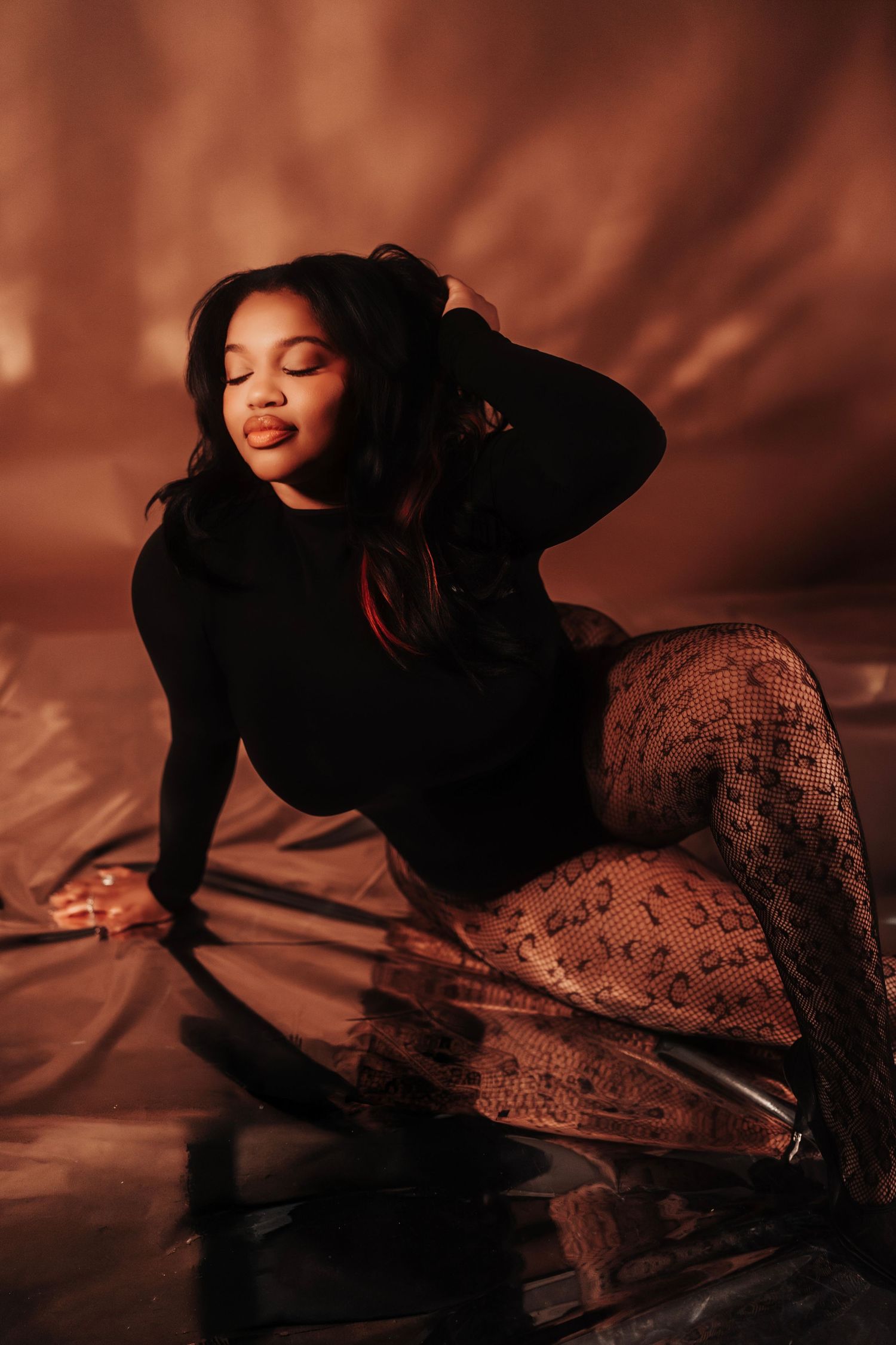 Woman in black top and patterned tights posing on reflective floor with warm brown backdrop.