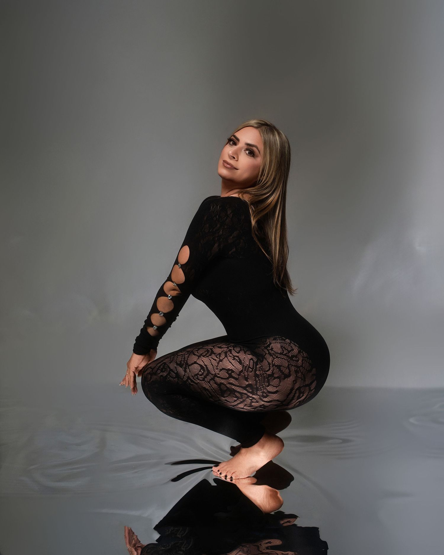 Woman in black lace bodysuit kneeling on reflective floor against neutral gray background.