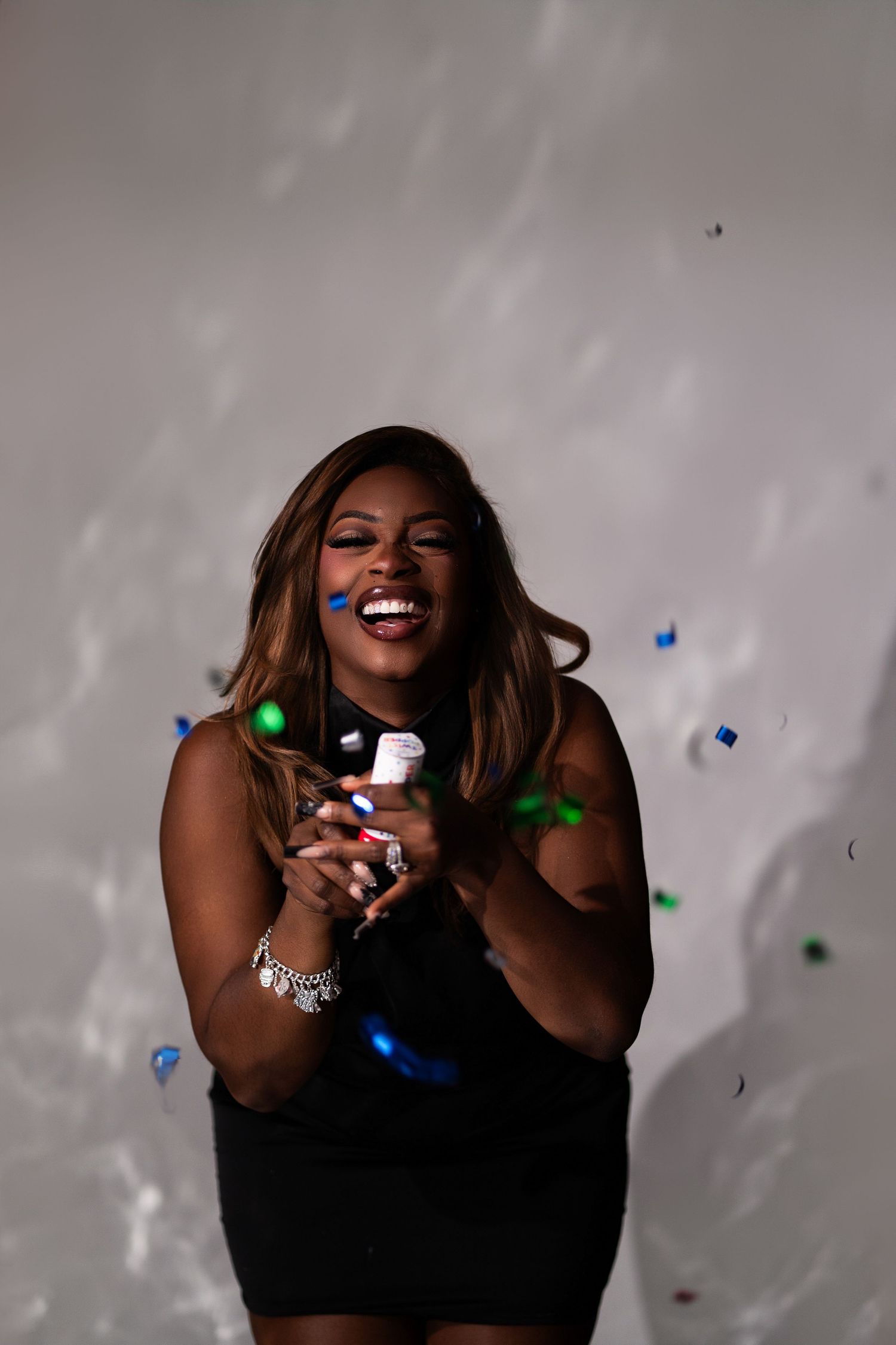 Joyful woman in black dress laughs as colorful confetti falls around her in studio.