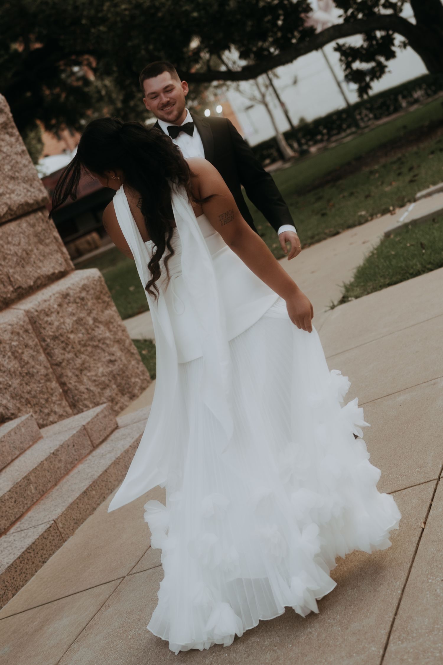 Smiling groom in black tuxedo embraces bride in flowing white ruffled wedding gown on decorative stone pathway.
