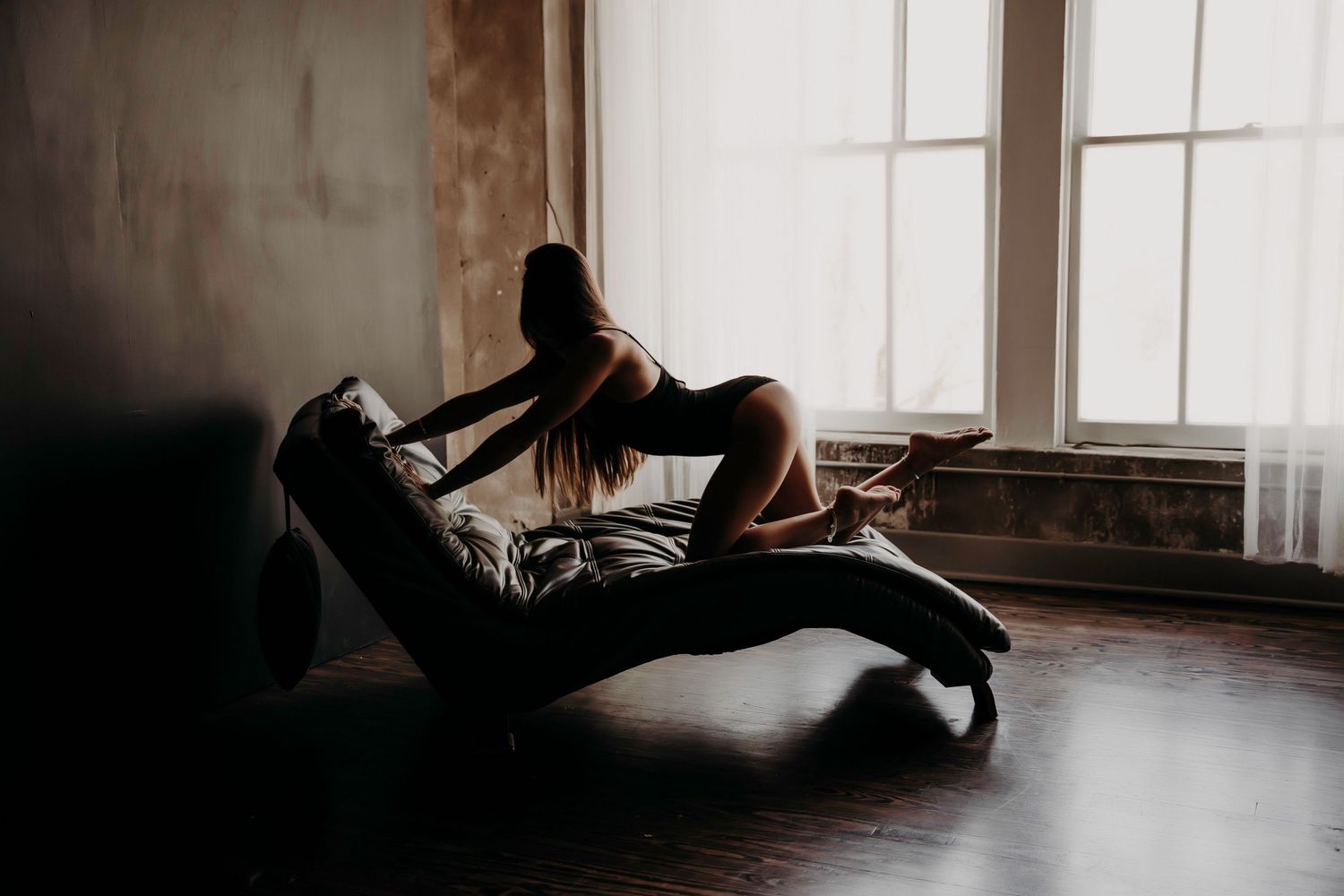 Silhouette of woman in lingerie posing on chaise lounge by bright window.