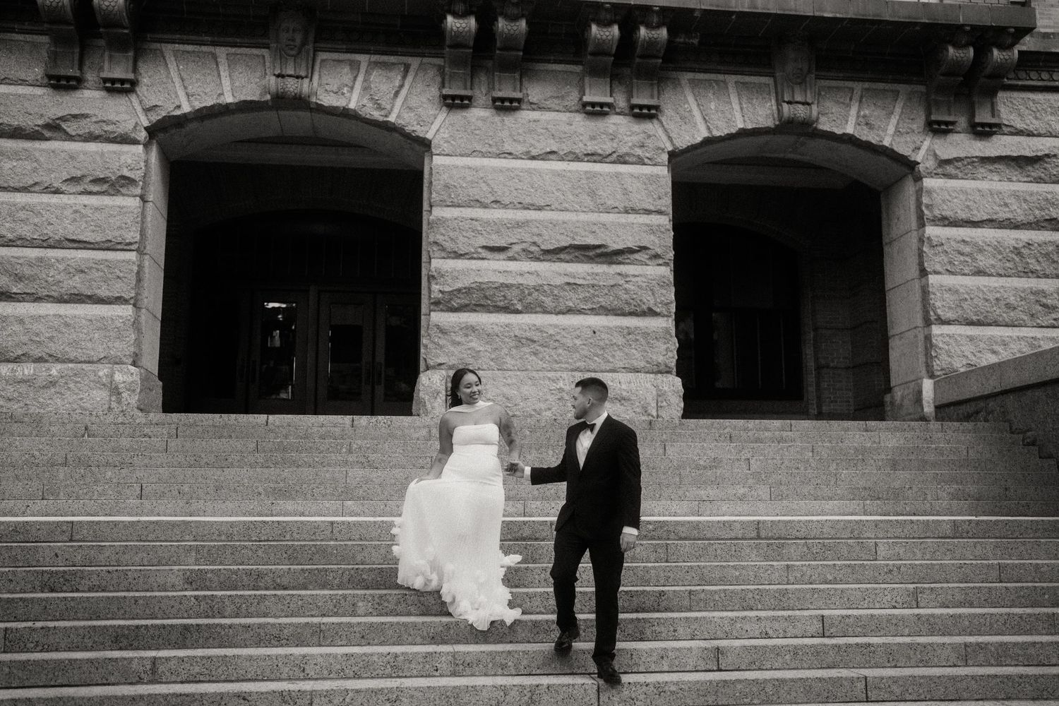 Black and white photo of bride and groom holding hands on wide stone staircase.