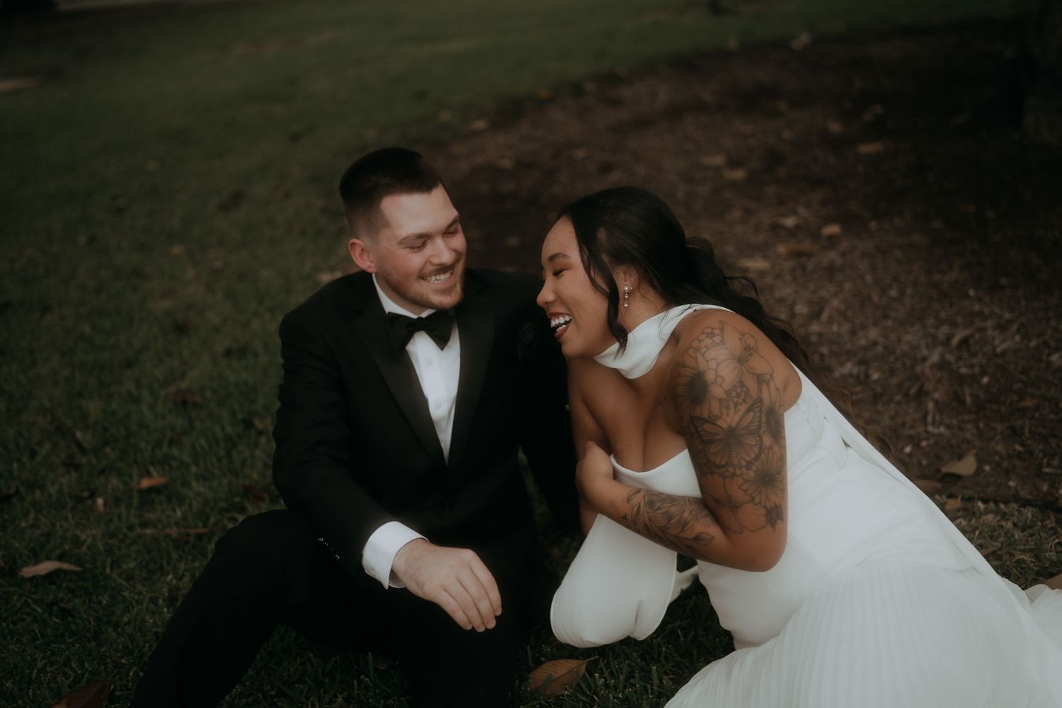 Bride and groom sit on grass laughing together during relaxed outdoor wedding photo session.