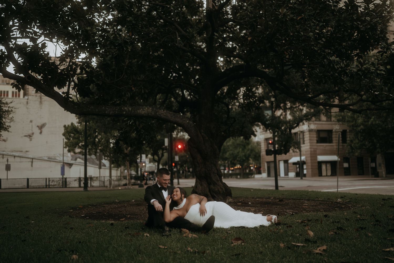 Bride and groom relax together under a large tree in an urban park during their wedding day.