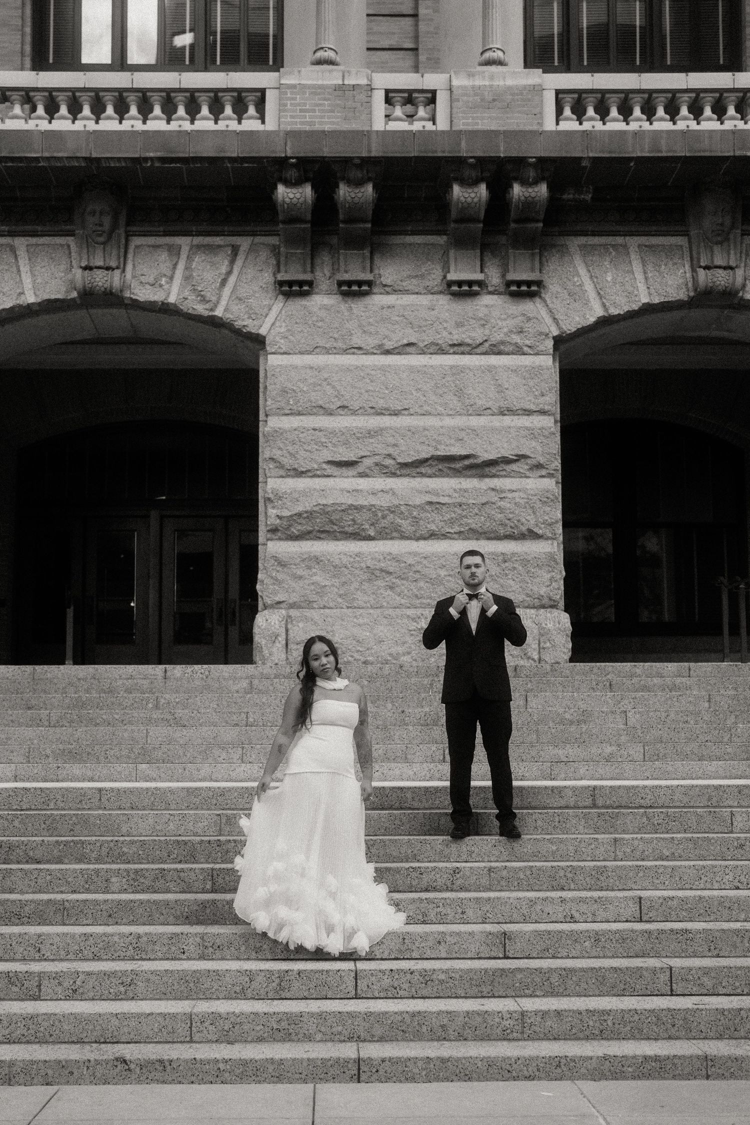 Black and white wedding portrait of bride and groom posing on grand steps of a classic stone building.