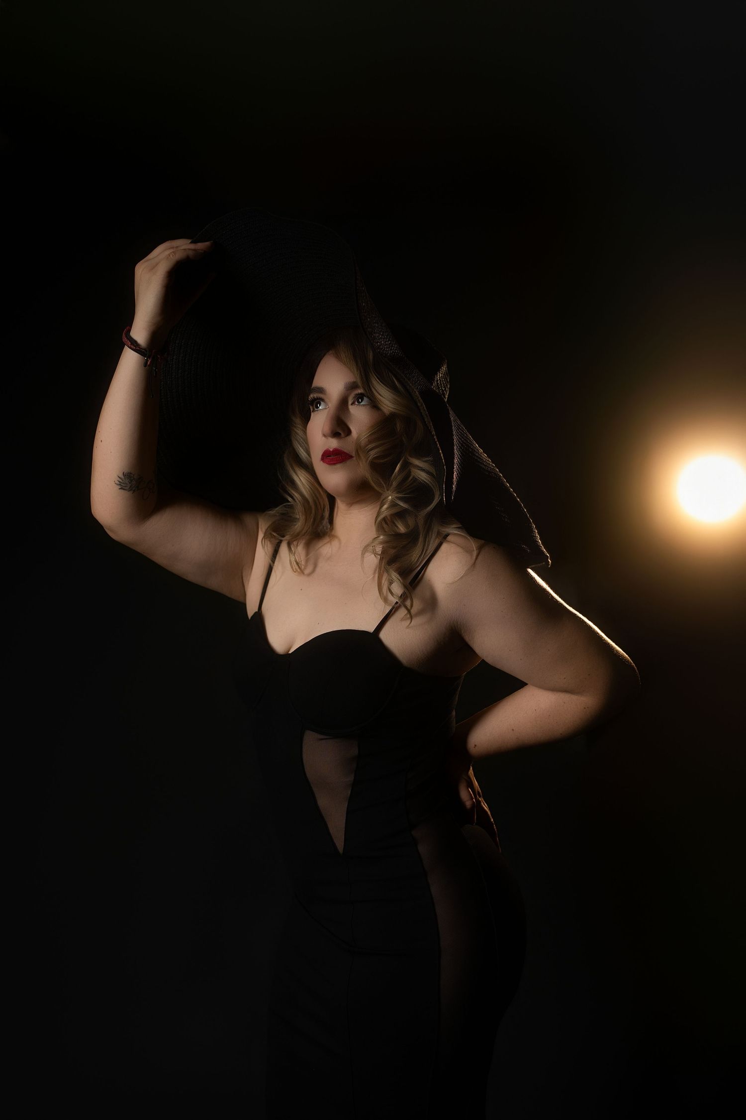 Woman in black corset dress holds hat while posing against dark background with warm backlight.