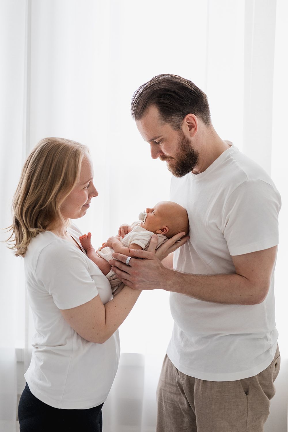 A mother and a father holding a newborn baby in front of a window.