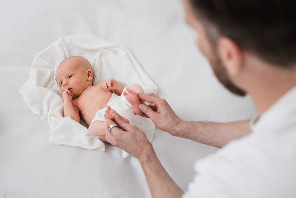 A newborn baby lays on a white surface and a father is holding the babysitter feet.