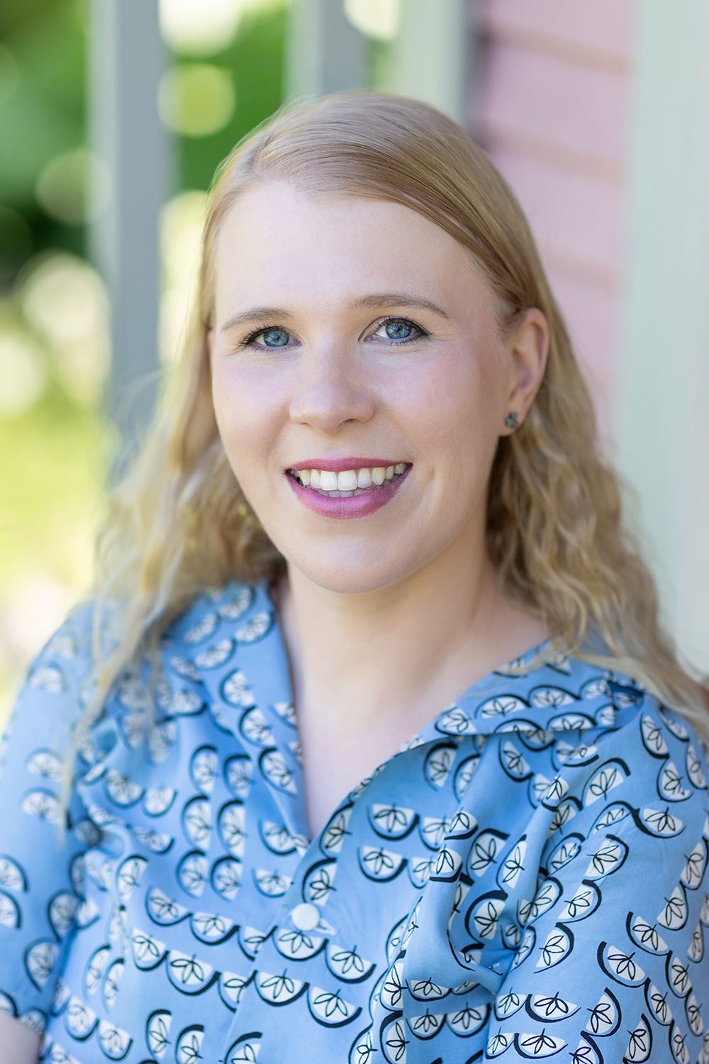 A blond hair woman smiling and looking in to a camera. Headshot. Outdoors.