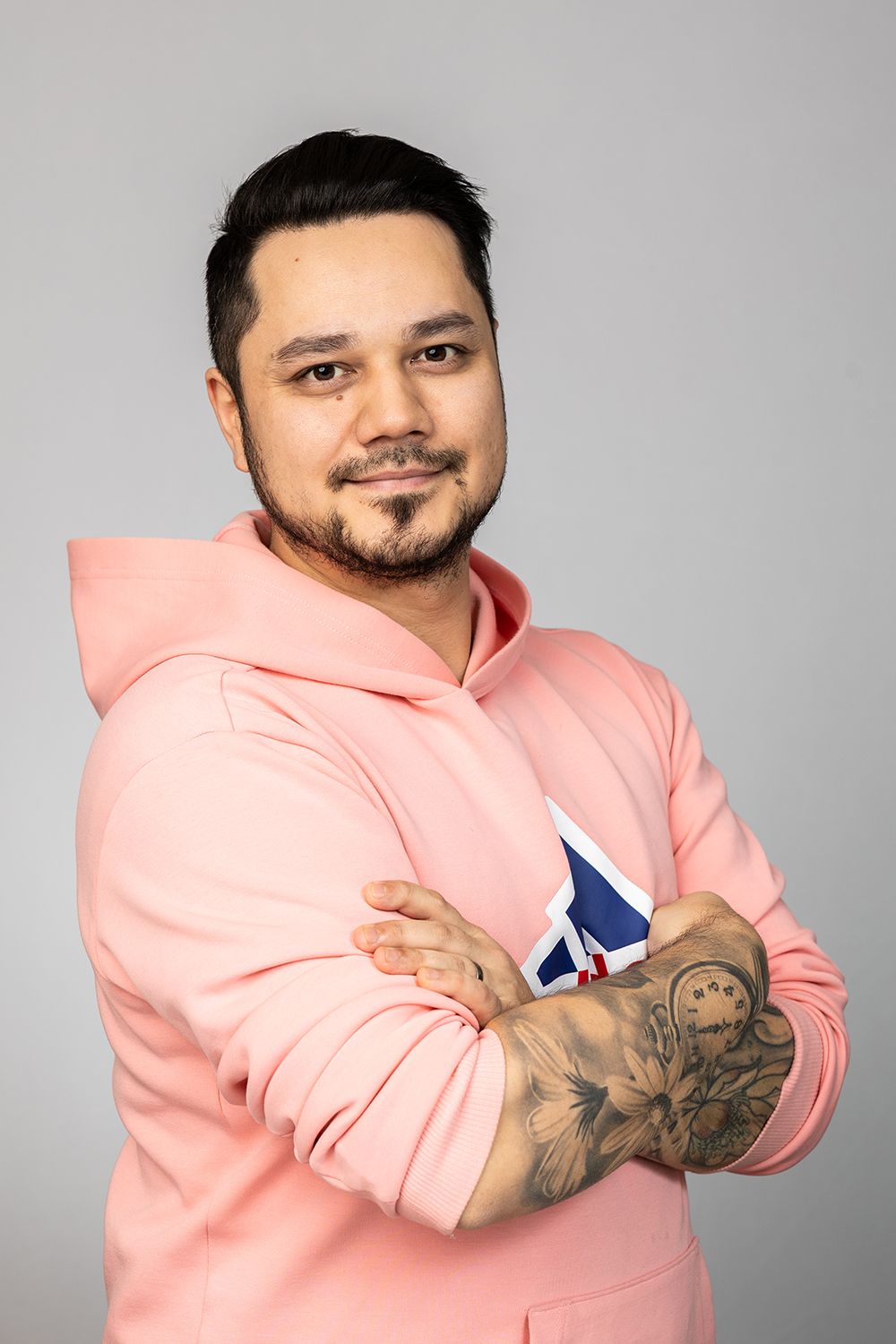 A dark hair man in a peach colored hoodie smiling and posing to a camera.