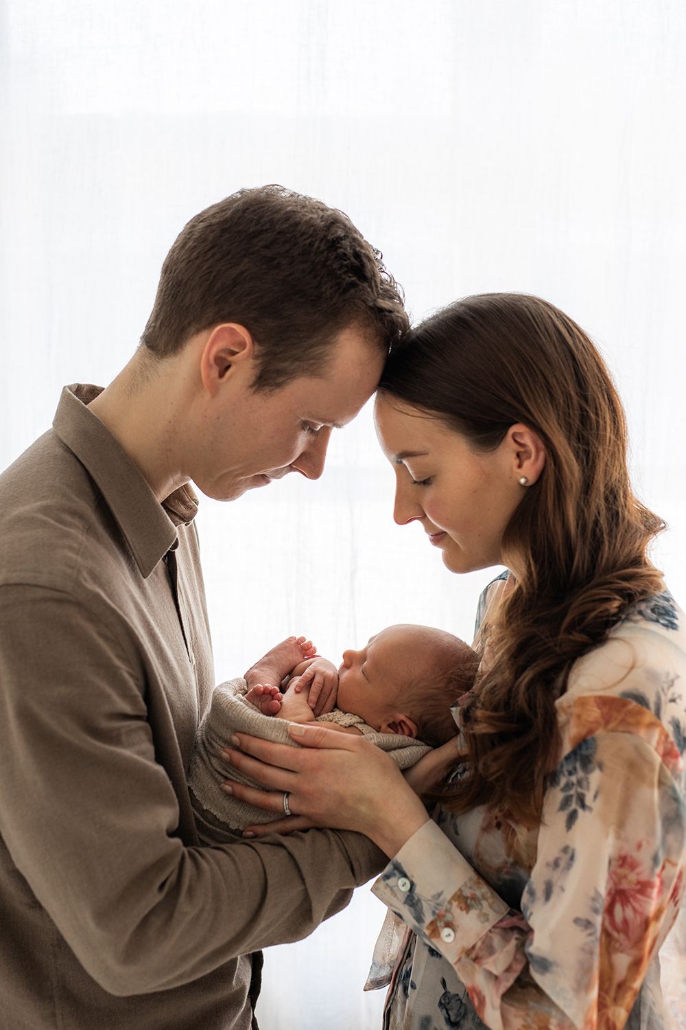 A mother and a father holding a newborn baby in front of a window.