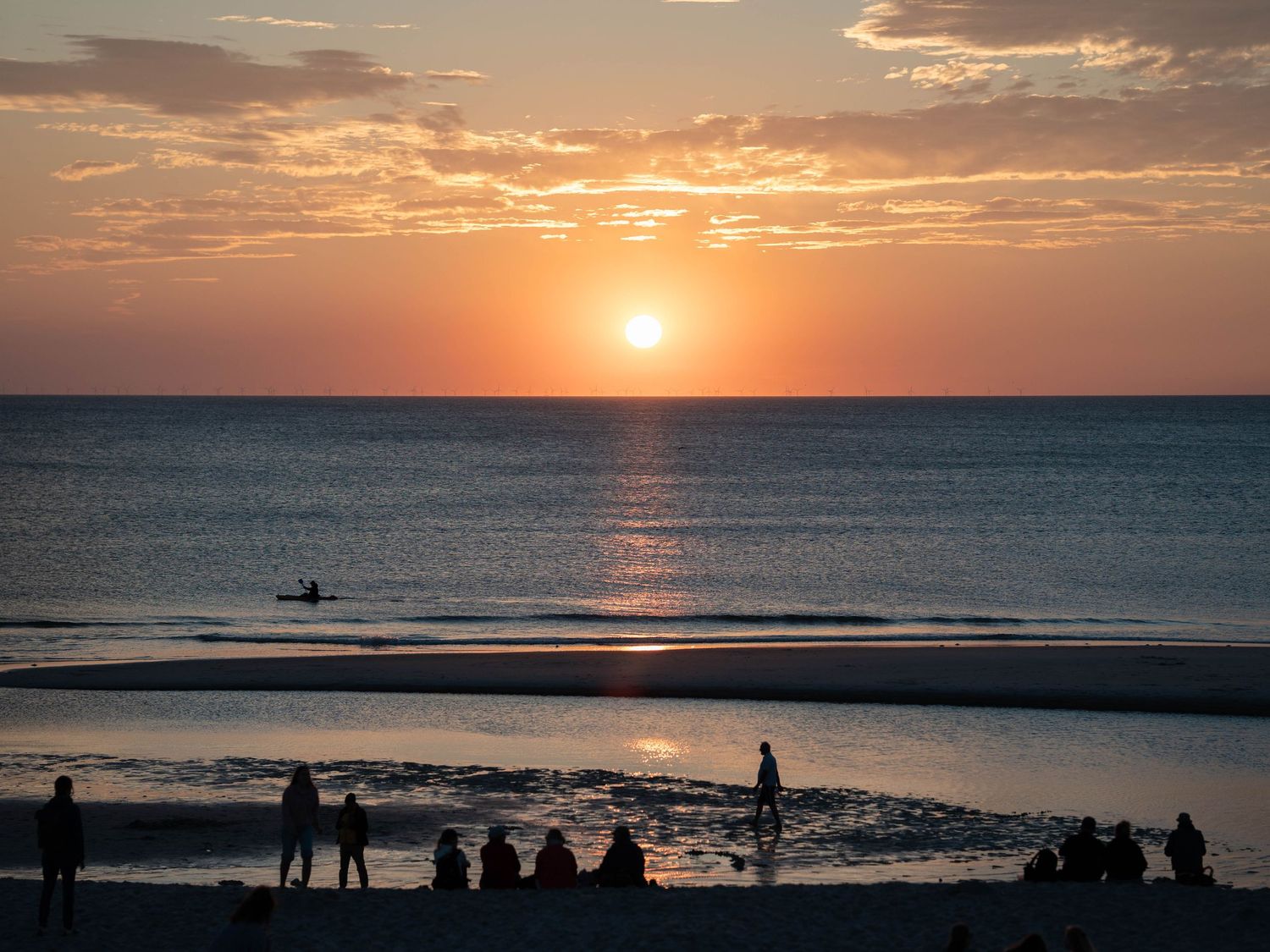 Stunning beach sunset with silhouetted people gathered on shore, golden sun reflecting on calm ocean waters.