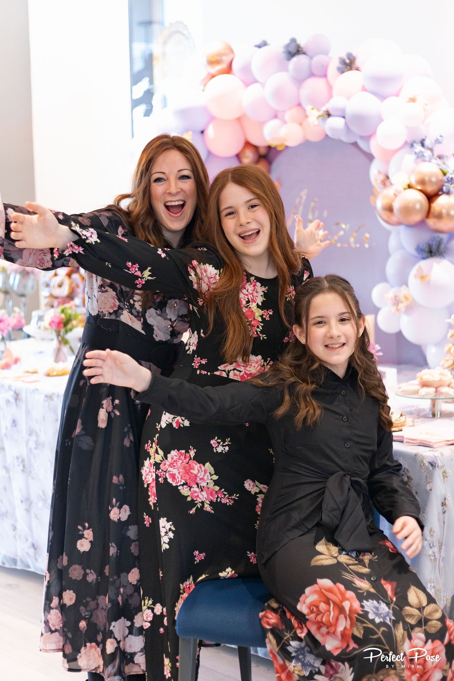 A woman and two girls in matching floral dresses smile joyfully at a party with a balloon arch backdrop.