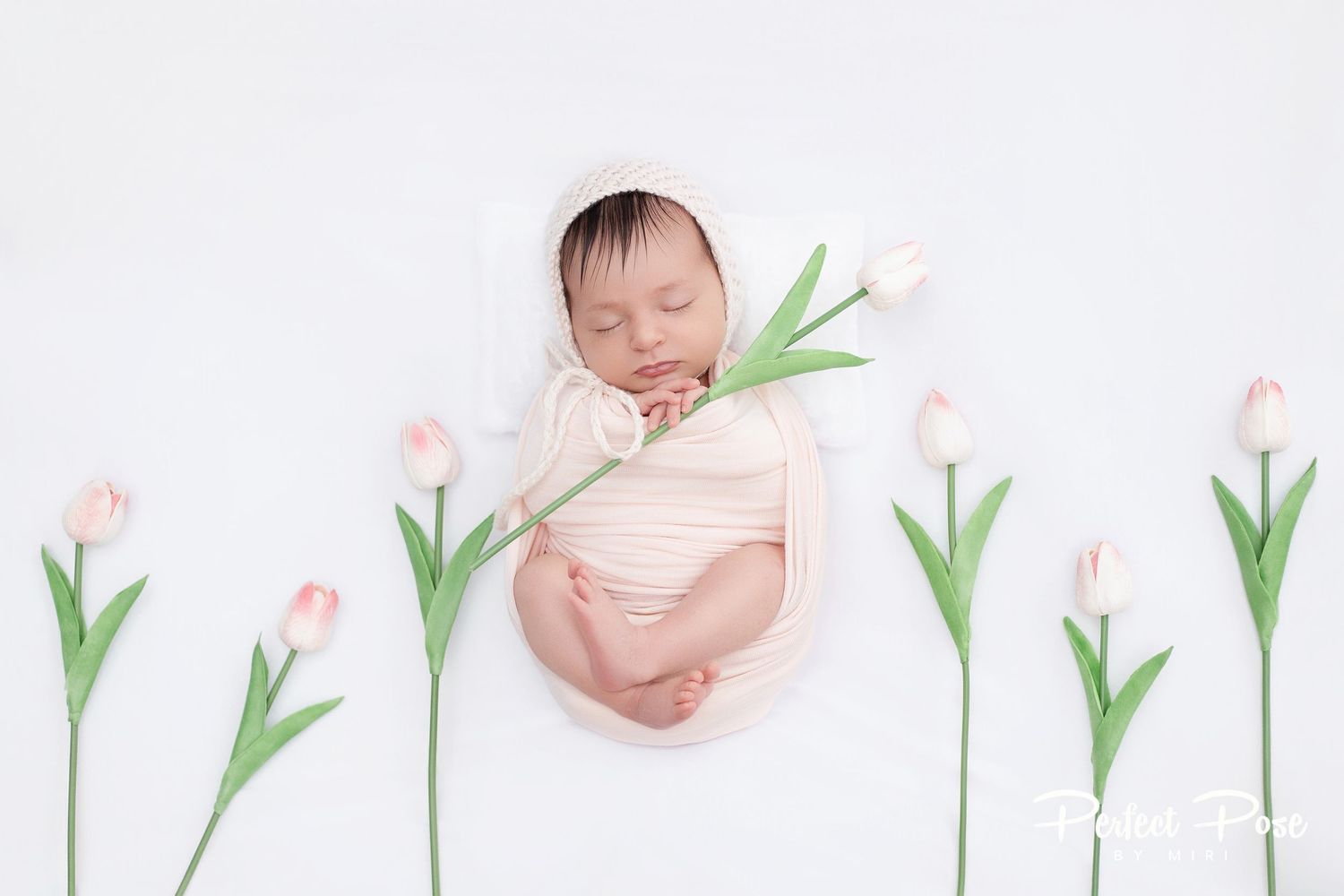 Sleeping newborn baby wrapped in pink swaddle surrounded by delicate pink tulips on white background.