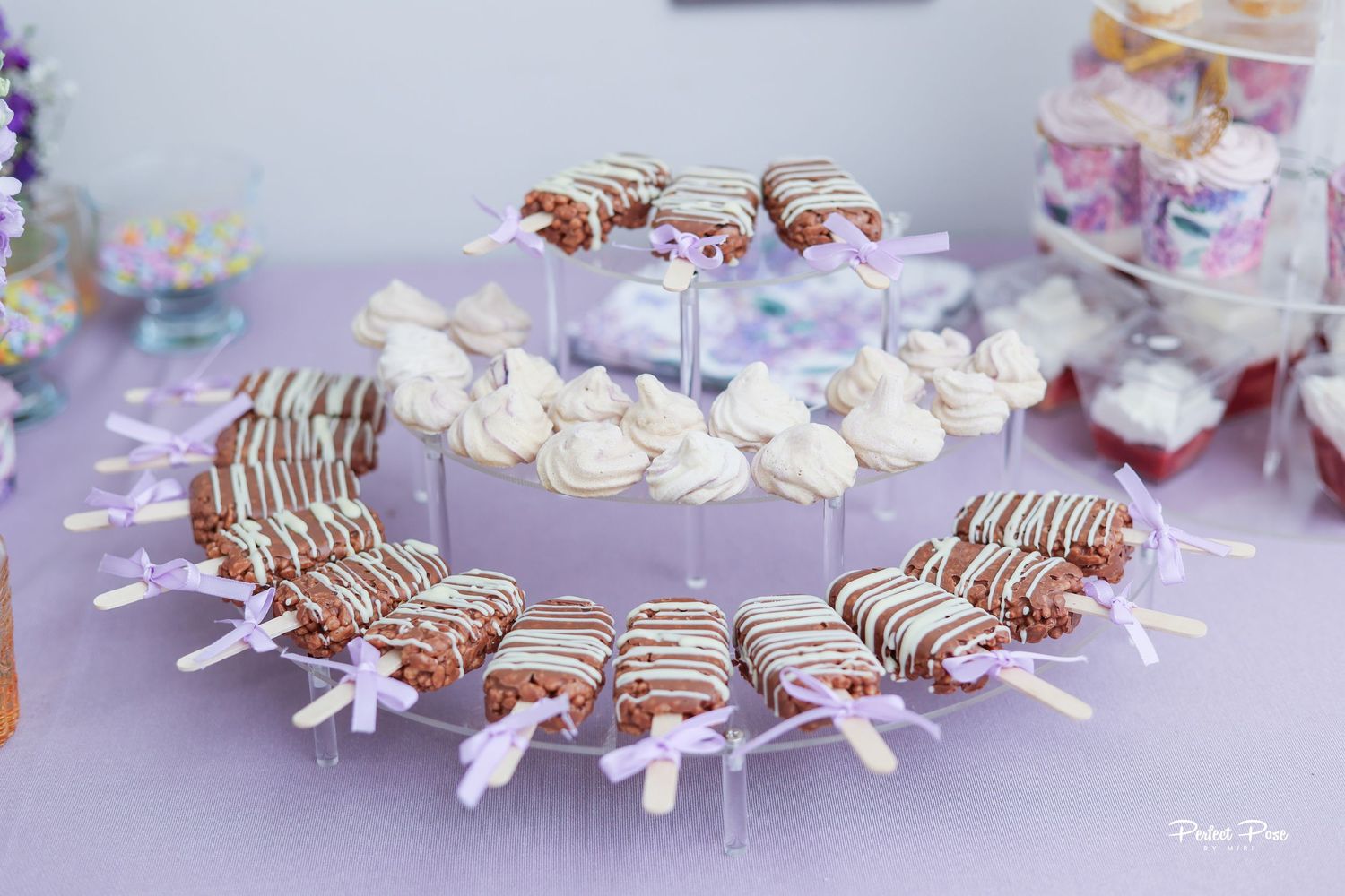 A tiered dessert display with chocolate-dipped cake pops and cream-topped treats decorated with purple ribbons.